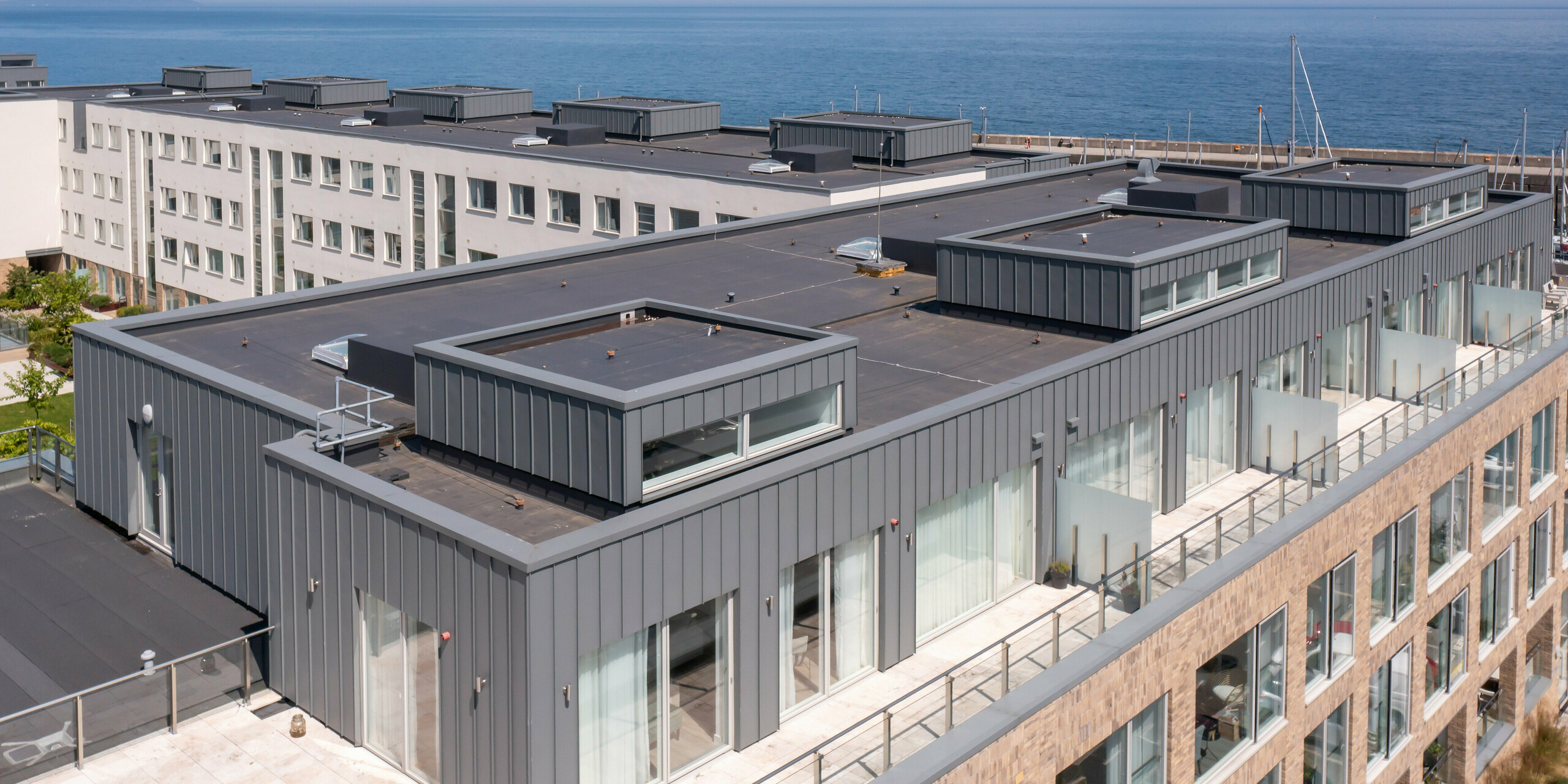 Marina Village Greystones in Wicklow, Ireland - Bird's eye view of the top floor of one of the modern apartment blocks. The picture shows a detailed view of the uninhabited roof terraces. The bright blue waters of the marina can be seen in the background. The floor is clad with high quality PREFALZ in P.10 light grey. The robust aluminium cladding provides durable protection and an aesthetic appearance. Large glass windows offer the future residents a breathtaking view of the sea and the marina. The perspective deliberately shows the impressive architecture against the picturesque backdrop of the marina.