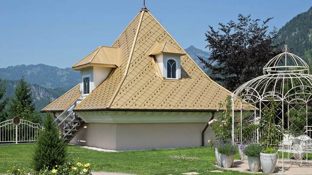 Kleiner Zubau eines Einfamilienhauses in Tirol mit PREFA Dachrauten in Mayagold und einem malerischen Garten im Vordergrund, umgeben von den Alpen.