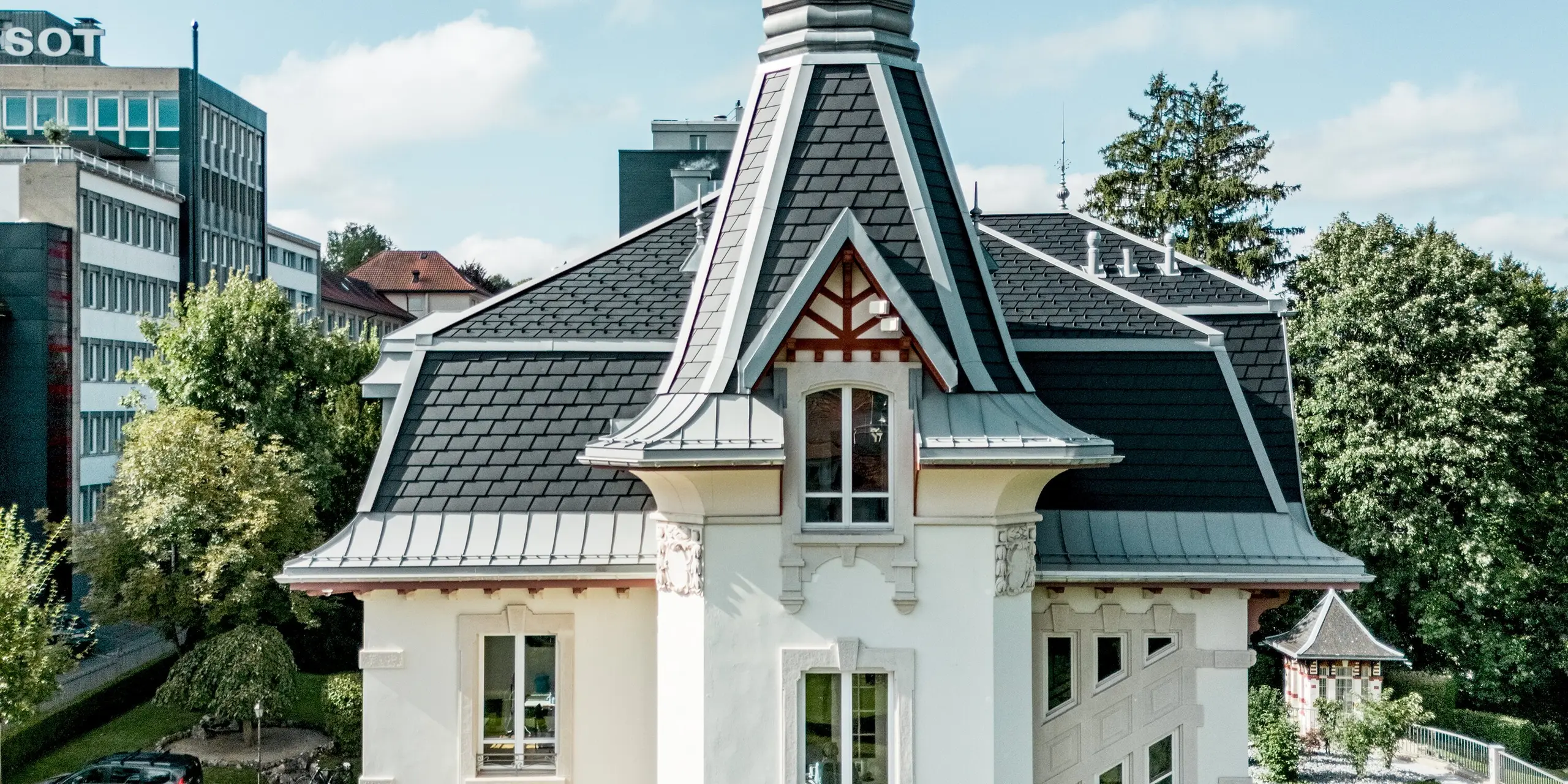 Markante Seitenansicht der Manufacture Horlogère du Locle in der Schweiz mit ikonischem Turm als Eyecatcher, der elegant in den Himmel ragt. Die komplexe Dachlandschaft fasziniert durch steil geneigte Flächen und präzise gegliederte Geometrie aus PREFA Dachschindeln in P.10 Schwarz. PREFALZ in Patinagrau setzt geschwungene Übergänge und feine Details perfekt um. Das langlebige, leichte Aluminiumdach überzeugt durch hohe Widerstandsfähigkeit, maximale Gestaltungsfreiheit und schont gleichzeitig die historische Bausubstanz nachhaltig.