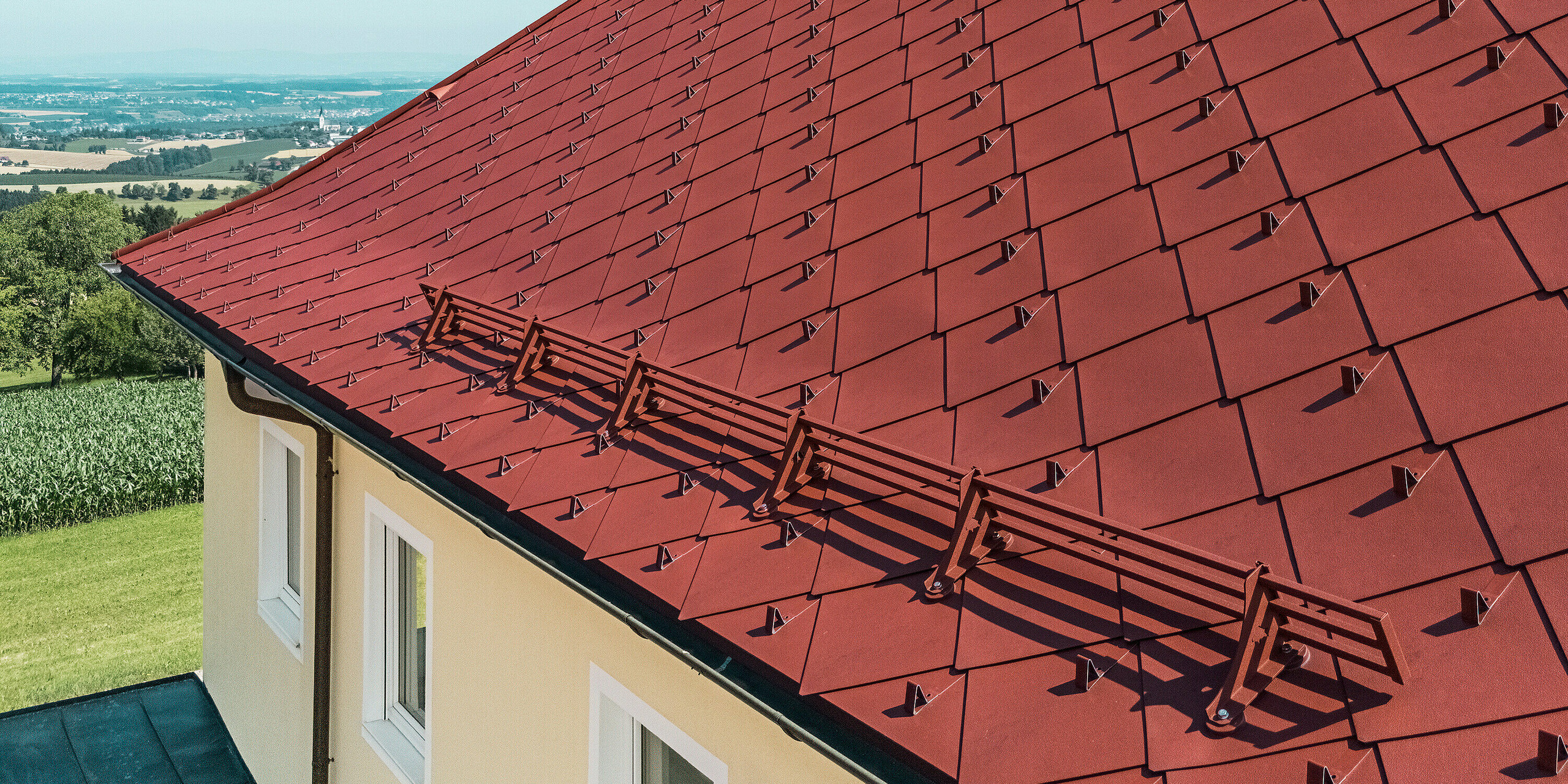 Das Bild zeigt das Dach eines sanierten Einfamilienhauses in Aschach an der Steyr, das mit der PREFA Dachraute 44 x 44 in der Farbe P.10 Oxydrot ausgestattet ist. Die markante rote Farbe des Daches fügt sich harmonisch in die umgebende Natur ein. Besonders auffällig ist der Schneefang aus Metall, der für Dach-Sicherheit sorgt. Zusätzlich wurden die einzelnen Dachrauten mit Schneestoppern ausgestattet, um das Abrutschen von Dachlawinen zu verhindern.