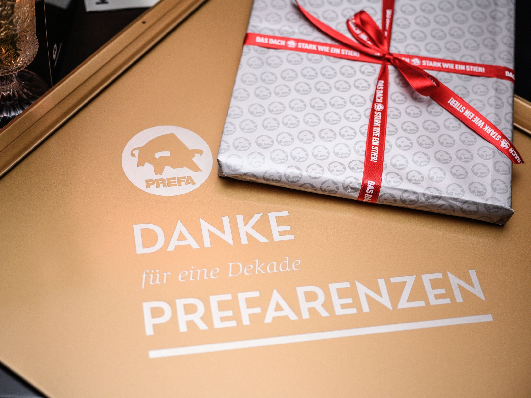A gift placed on a golden surface featuring the PREFA logo and the inscription 'THANK YOU for a decade of PREFARENCE'. The gift is wrapped in silver paper adorned with small PREFA logos and tied with a red ribbon that reads 'STRONG AS A BULL'