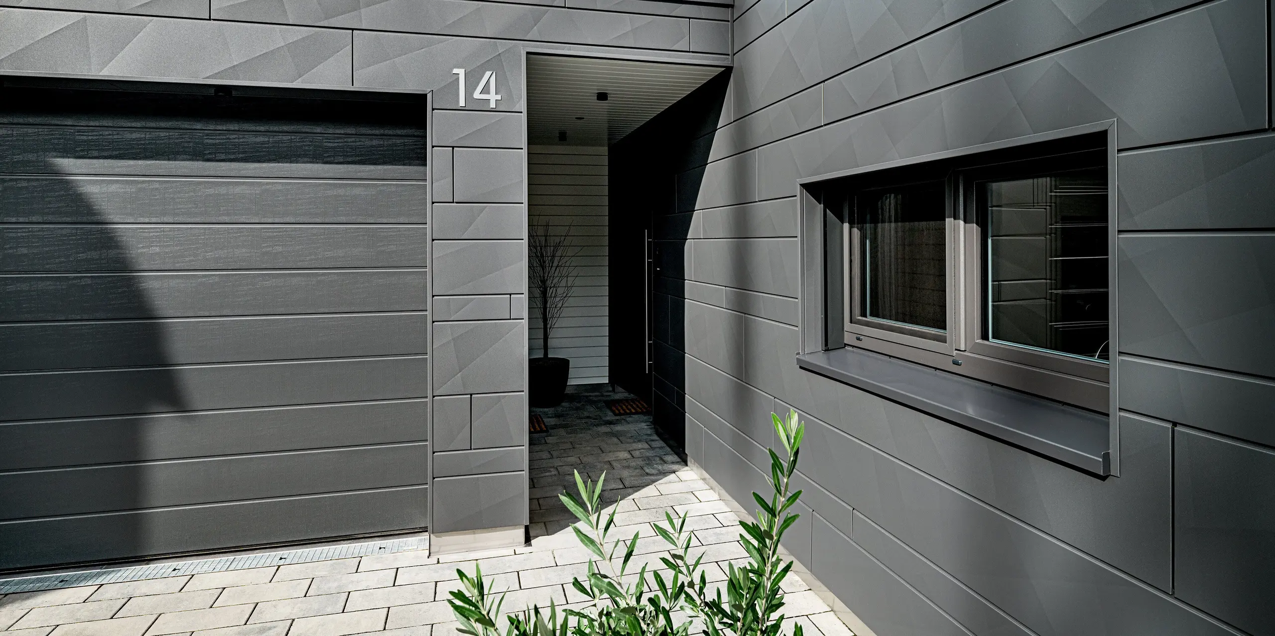 Eingangs- und Garagenbereich eines modernen Einfamilienhauses in Heidenheim mit vollständig verkleideter PREFA Siding.X Aluminiumfassade in P.10 Dunkelgrau. Die markanten Kantstrukturen der Paneele erzeugen ein lebendiges, geometrisches Muster. Die Hausnummer „14“ steht klar sichtbar auf dem Fassadenelement über dem überdachten Zugang. Rechts prägt ein großes, präzise eingefasstes Fenster die Szene, während die gepflasterte Zufahrt und dezente Bepflanzung den modernen Gesamteindruck abrunden. Die langlebige PREFA Oberfläche bietet hohen Schutz und zeitlose Stabilität.