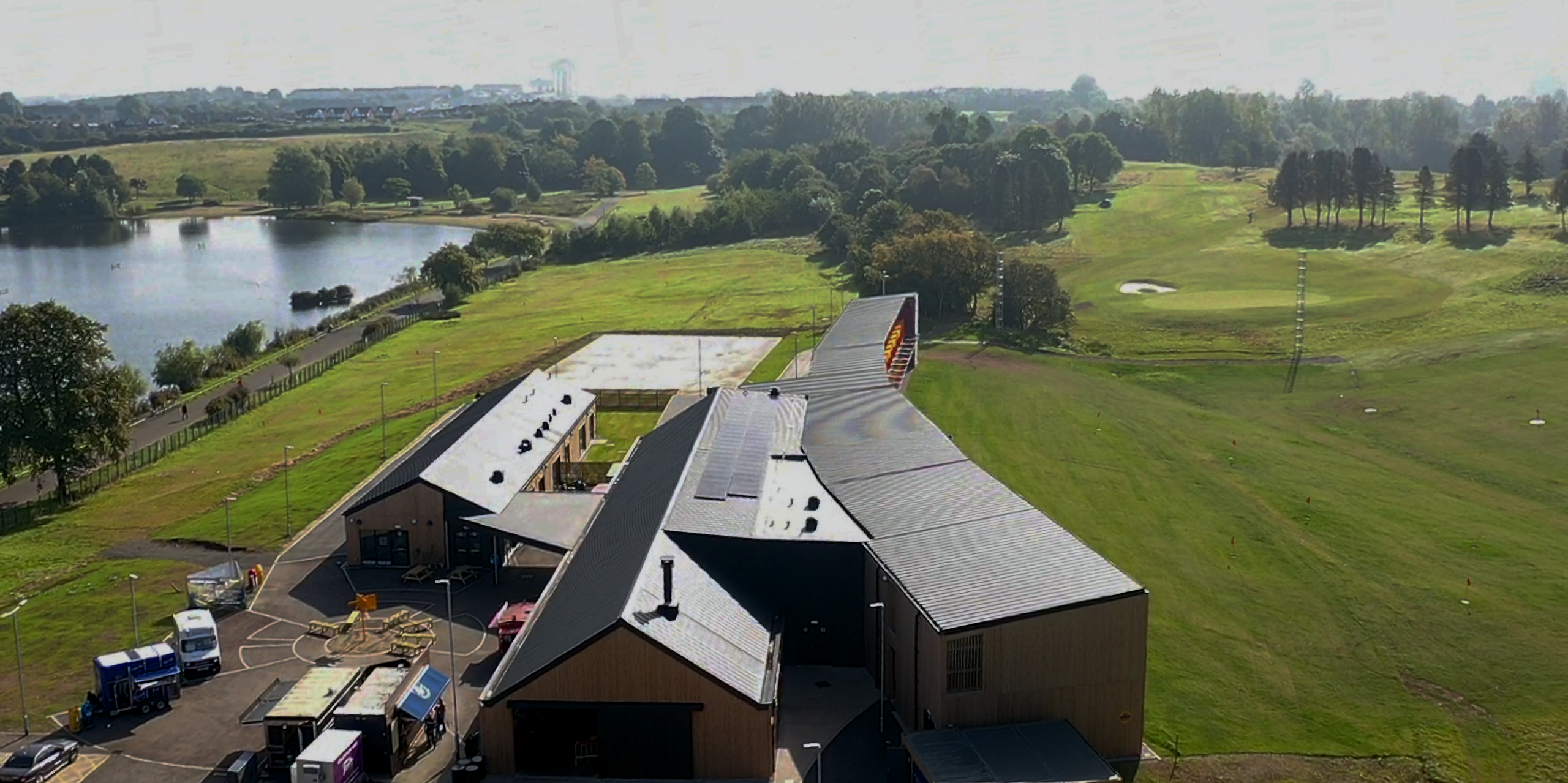 Aerial view of the 'Golf it! golf course in Glasgow, which is set in a stunning landscape with a lake and an extensive golf course. The buildings are clad with PREFALZ in P.10 Black. This view demonstrates the adaptability and aesthetic quality of PREFA aluminium products in a natural environment.