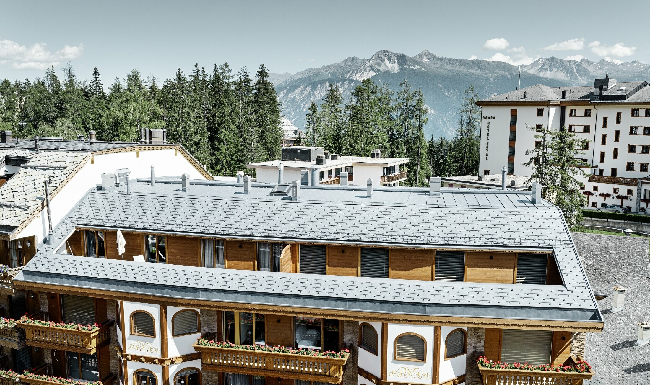 Apartment building in Crans-Montana (Switzerland) with mountains in the background, a façade with lovely wooden elements and a PREFA aluminium shingle roof in stone grey