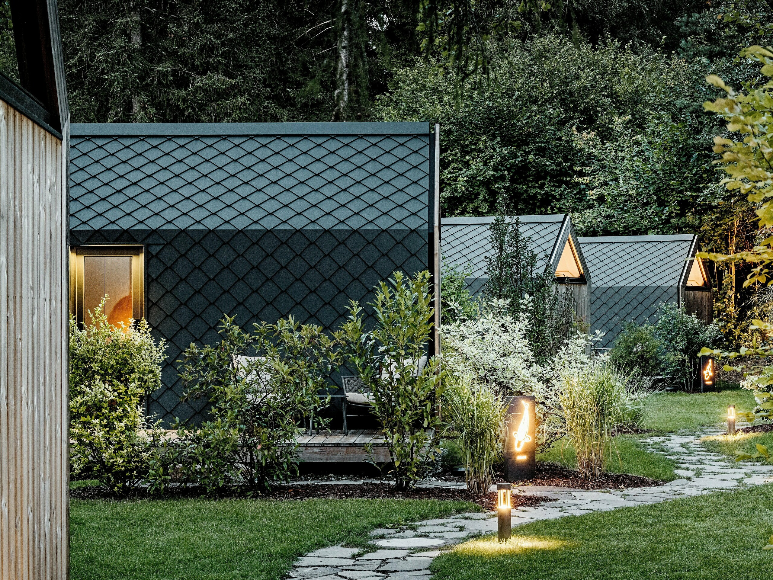 Ein beleuchteter Gartenweg führt durch das Glamping‑Resort Secret Koi Garden am Natterer See zu mehreren minimalistischen Tiny Houses. Die kompakten Baukörper sind vollständig mit PREFA Dach‑ und Wandrauten aus Aluminium in P.10 Anthrazit verkleidet, deren fischhautartige Struktur im Abendlicht elegant wirkt. Eingebettet in üppiges Grün verbinden die langlebigen, witterungsbeständigen PREFA Aluminiumoberflächen hochwertige Architektur mit Natur, Ruhe und nachhaltigem Design.