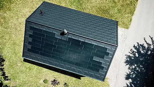 Die spektakuläre Drohnenaufnahme zeigt das PREFA Solardach von Haus Immi aus der Vogelperspektive. Schwarze Solardachplatten gehen nahezu unsichtbar in die Dachplatten R.16 in P.10 Schwarz über und formen eine durchgängige Dachfläche. Das integrierte Aluminiumsystem vereint nachhaltige Solarstromerzeugung, hohe Widerstandsfähigkeit und klare Architektur ohne optisch aufgesetzte Photovoltaikanlage im alpinen Raum.