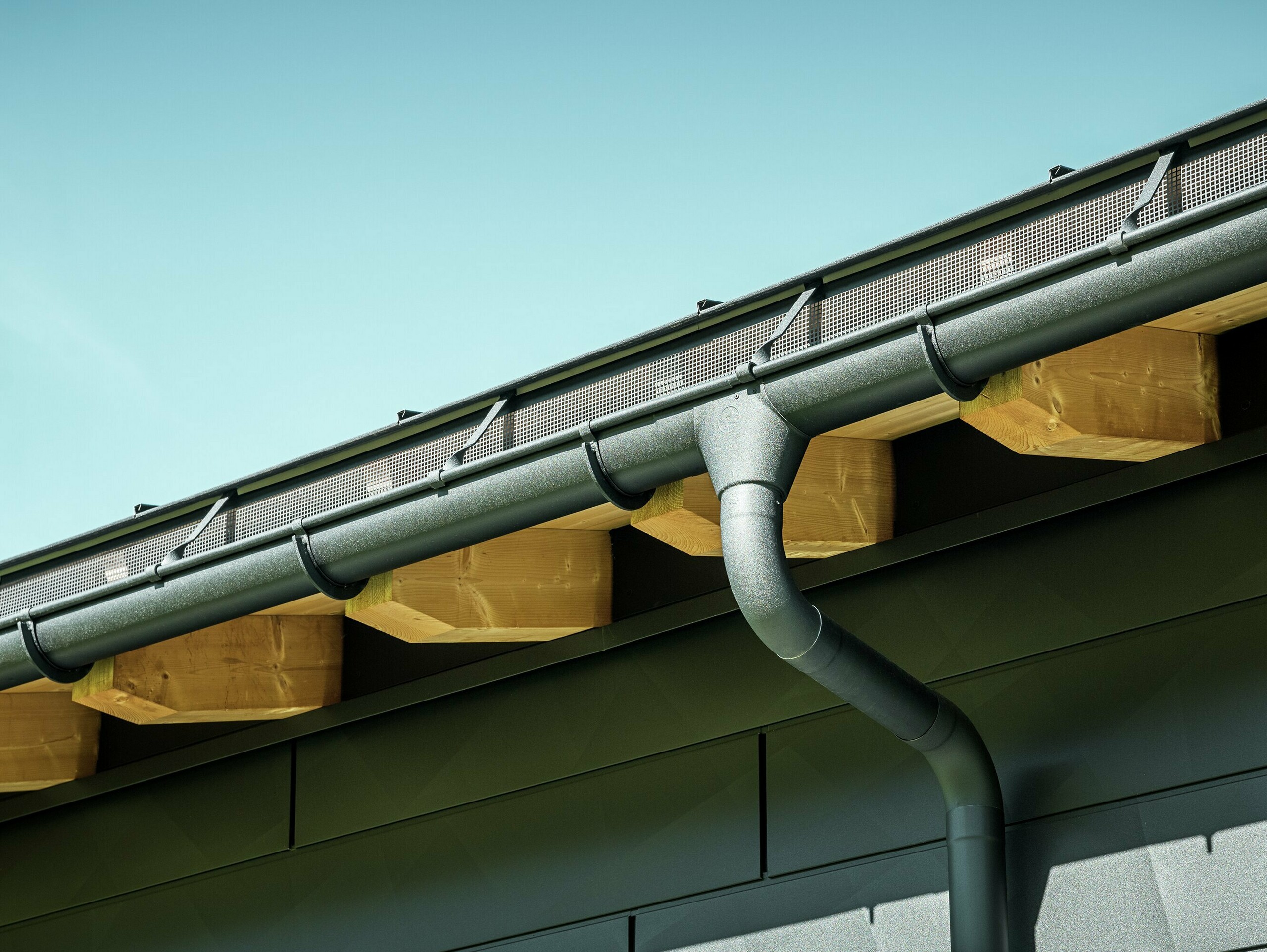 PREFA half-round gutter with gutter outlet and reinforcement hanger in P.10 black Close-up of the façade of a modern detached house in Ascharina, Switzerland. The sides of the building were clad with striking PREFA Siding.X façade panels in P.10 black. The PREFA roof drainage system with half-round gutter and downpipe is colour-coordinated.