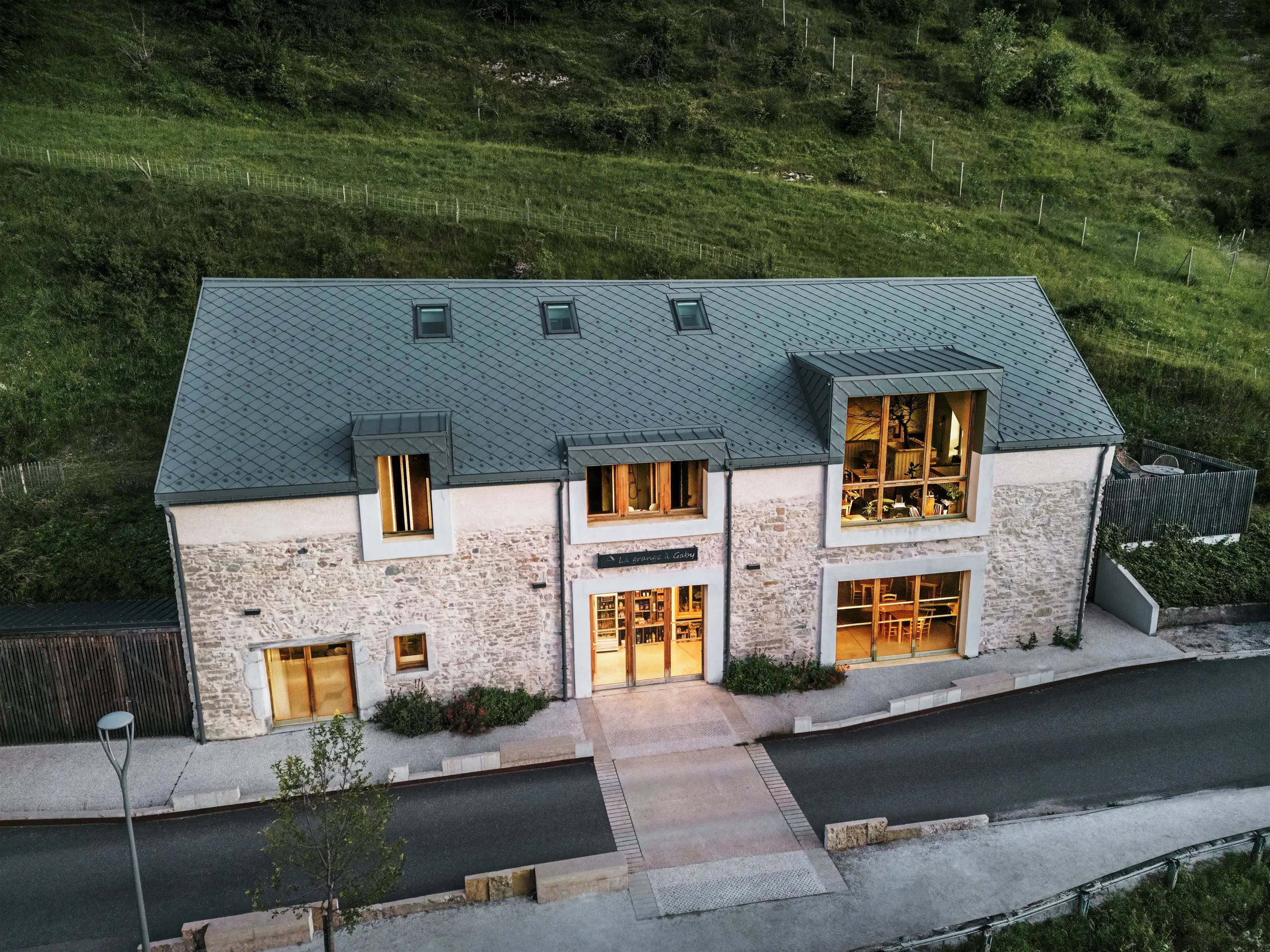 Evening view of the restored barn ‘La Grange à Gaby’ in Engins, France. The building combines traditional stone masonry with a modern PREFA aluminium roof made of 29 × 29 cm rhomboid tiles. The large windows and the illuminated interior emphasise the welcoming atmosphere of the new cultural centre.
