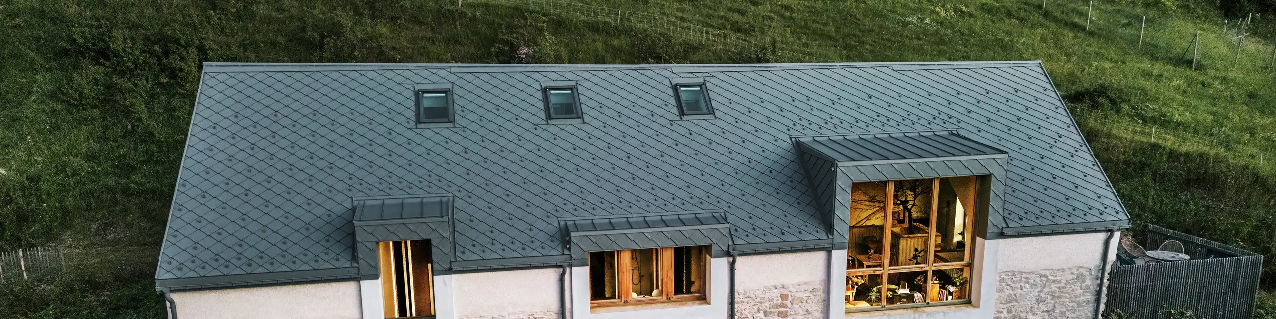 Evening view of the restored barn ‘La Grange à Gaby’ in Engins, France. The building combines traditional stone masonry with a modern PREFA aluminium roof made of 29 × 29 cm rhomboid tiles. The large windows and the illuminated interior emphasise the welcoming atmosphere of the new cultural centre.