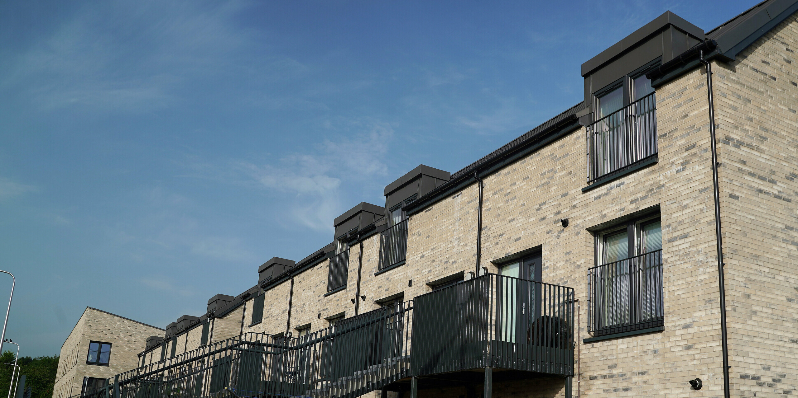 View of a modern, multi-storey residential complex in Edinburgh with a roof area of around 1000 square metres, covered with PREFALZ in P.10 dark grey. The characteristic dark grey colouring of the roof forms an elegant contrast to the light brick façade. The staircases and drainage pipes for the rainwater are also in dark grey tones. A red sign indicates that parking is prohibited and requires 24-hour access.