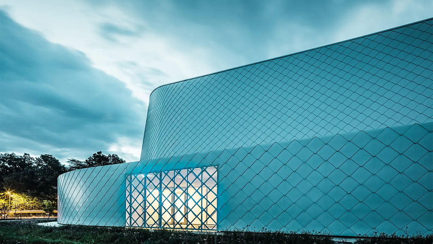 Lateral evening evening shot of the illuminated building, the window is barred with the same patterns as that of the rhomboid tile façade.