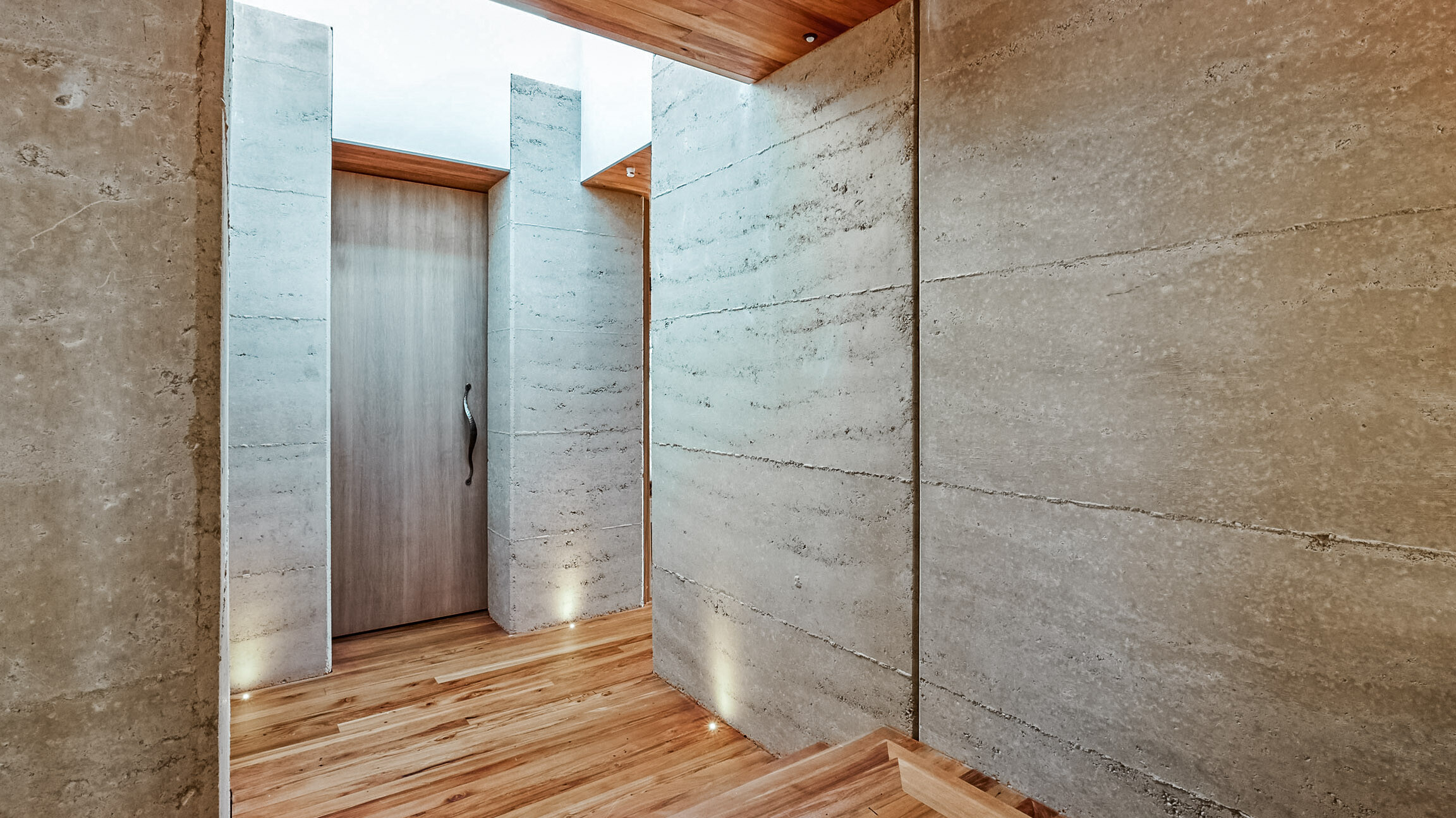 A shot of the interior: The wooden floor with its floor lights, a wooden door and the walls made of rammed earth are visible.