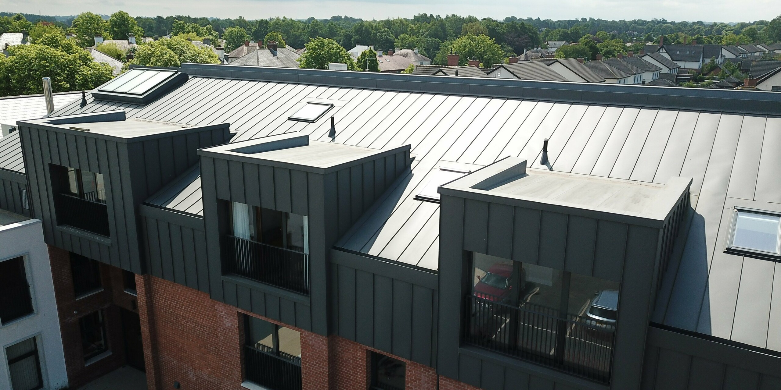 A bird's eye view of the roof of Harberton Hall in Belfast, Northern Ireland. This modern building for supported living combines PREFALZ aluminium in P.10 dark grey with a classic tile façade. The flat roof dormers with their large windows ensure that the interior is flooded with light. The standing seam cladding creates a harmonious connection between contemporary design and traditional construction.