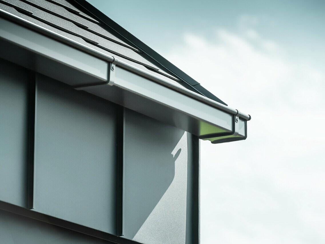 Corner of house with the PREFA box gutter in anthracite, fastened with the PREFA gutter brackets, while the façade is clad with PREFA Prefalz underneath.