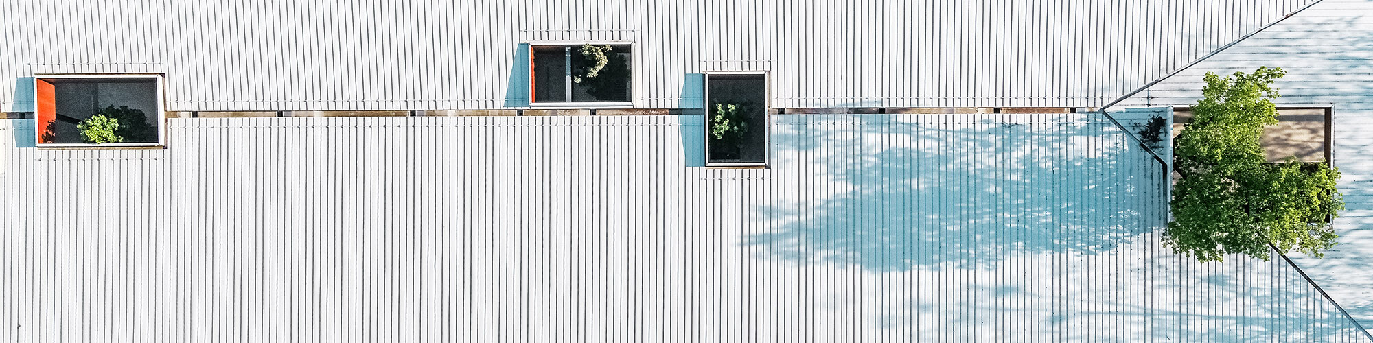 The picture shows an aerial view of the white Prefalz roof of the "Autobusni Kolodvor Slavonski Brod" bus stop in Croatia. The roof is interrupted by several rectangular openings through which green trees grow, creating a harmonious connection between the architecture and nature. The clean lines and white surface of the roof are interrupted by the shadows of the trees, creating an interesting pattern. The image highlights the innovative and environmentally friendly design of the station, which is characterised by its functionality and aesthetic integration into the surroundings.