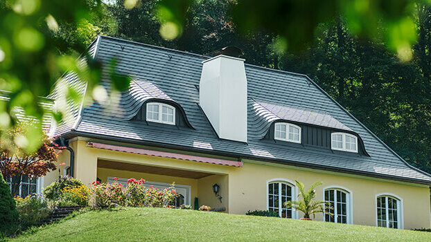 Detached house with a roof newly renovated with PREFA shingles in anthracite with rounded dormers (eyebrow dormers) and a white chimney.