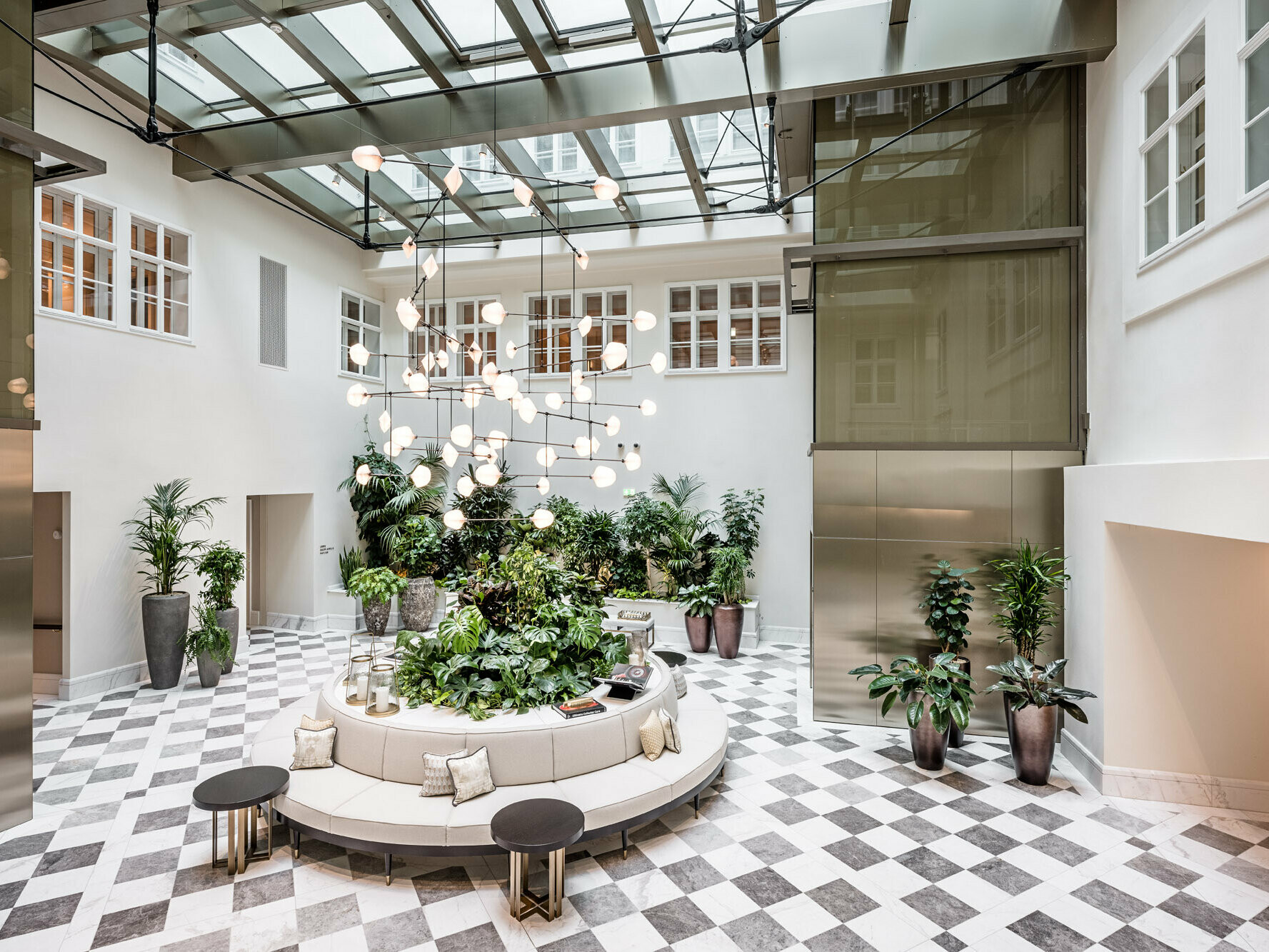 Interior of the luxury hotel, where the colours bronze, grey and white dominate: There are seating areas and larger potted plants on a marble floor, a modern lamp is hanging down into the room from the ceiling.