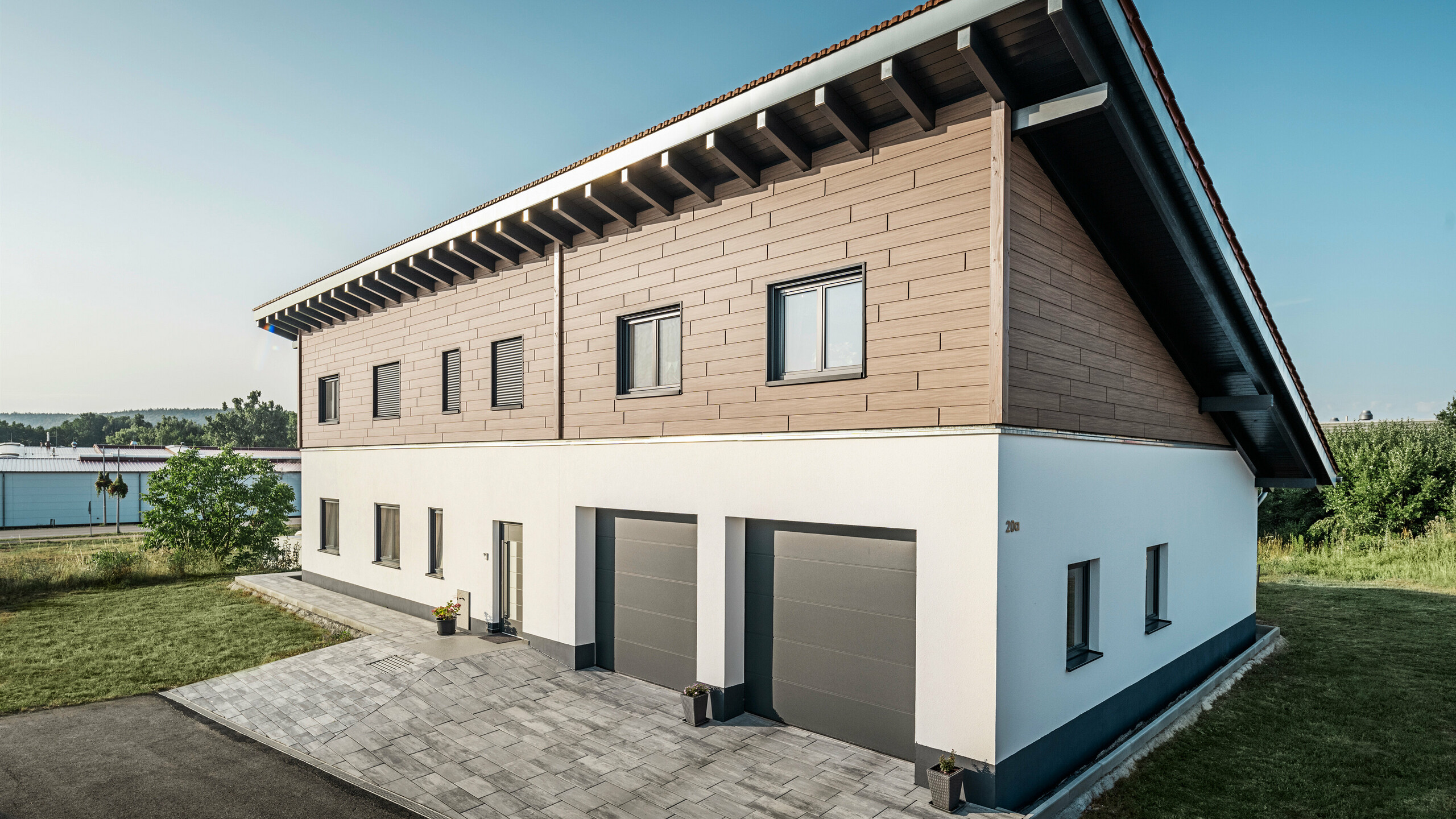 View from the side of a detached house with a monopitch roof in Bruck. The upper storey is clad with PREFA sidings in Walnut Brown. Two garage doors in anthracite can be seen on the right-hand side of the picture. The entrance door is located in the centre of the building. The plinth is in a plain white colour. 