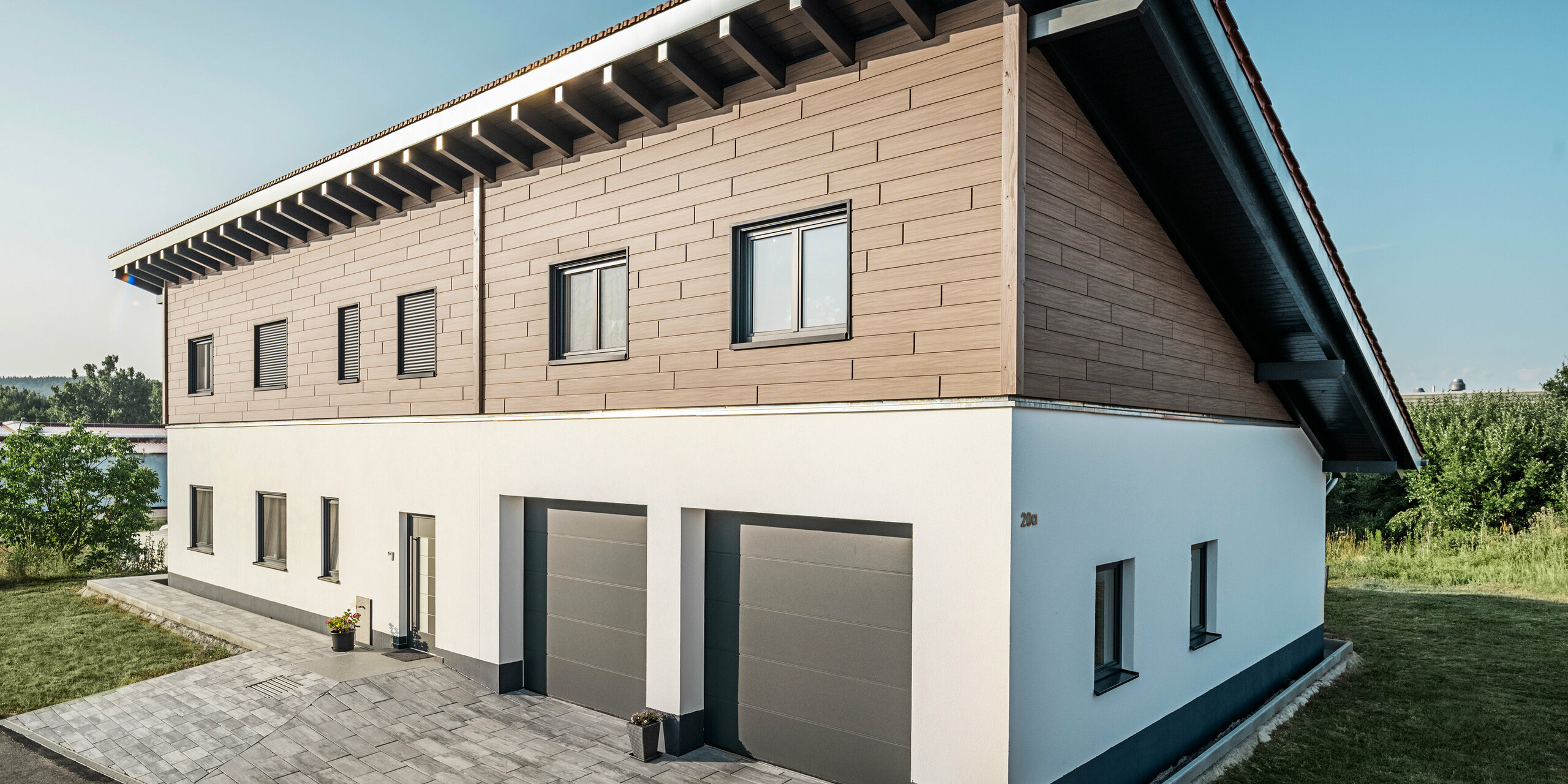 View from the side of a detached house with a monopitch roof in Bruck. The upper storey is clad with PREFA sidings in Walnut Brown. Two garage doors in anthracite can be seen on the right-hand side of the picture. The entrance door is located in the centre of the building. The plinth is in a plain white colour. 