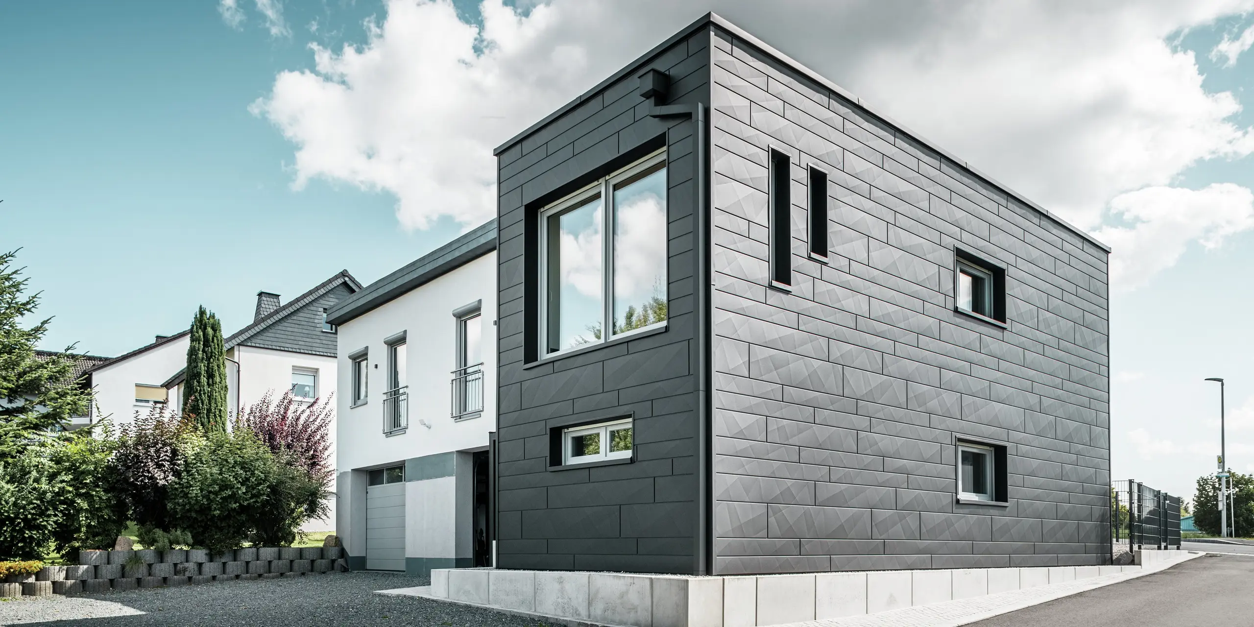 Den stilfulde og moderne facade på det renoverede enfamiliehus i Teuschnitz viser de klart strukturerede PREFA Siding.X-paneler i P.10 sort. Den geometriske placering af de højkvalitative pladepaneler giver bygningen en markant, tidløs æstetik. Vinduerne er indrammet af sorte aluminiumskompositplader, hvilket afrunder designet harmonisk. Det integrerede PREFA tagafvanding med kvadratrør sikrer en funktionel og æstetisk løsning.