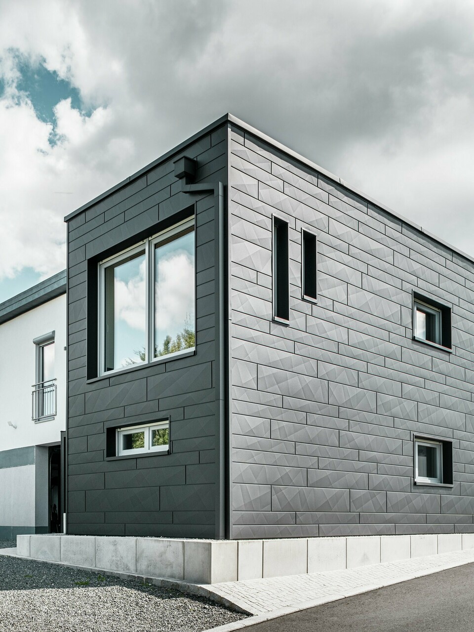 Die stilvolle und moderne Fassade des sanierten Einfamilienhauses in Teuschnitz zeigt die klar strukturierten PREFA Siding.X Paneele in P.10 Schwarz. Die geometrische Anordnung der hochwertigen Blechpaneele verleiht dem Gebäude eine markante, zeitlose Ästhetik. Die Fenster sind von schwarzen Aluminium Verbundplatten umrahmt, was das Design harmonisch abrundet. Die integrierte PREFA Dachentwässerung mit dem Quadratrohr sorgt für eine funktionale und ästhetische Lösung.