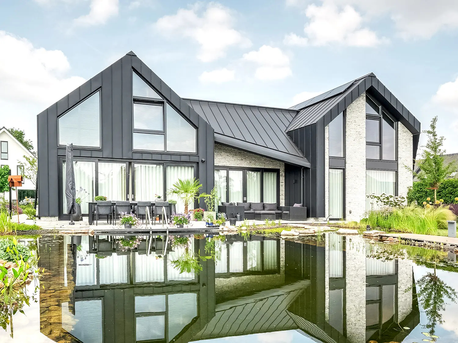 Modern detached house with characteristic, pointed gable roofs, covered with black Prefalz roof panels, reflected in a tranquil garden pond. The façade combines dark wall elements with light-coloured brick areas and large windows. A terrace with garden furniture invites you to linger and is harmoniously integrated into the lush, flowering garden landscape.