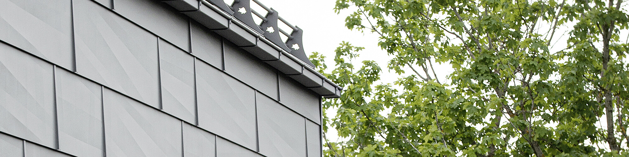Close-up of the light grey aluminium façade with the transition to the roof, framed by treetops in the right of the photo.