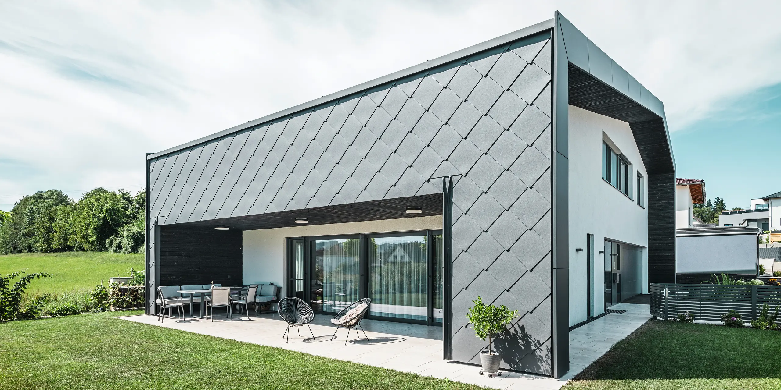 Well-kept garden area of a detached house in Aschach. The building is stylishly clad with dark grey PREFA aluminium rhomboid façade tiles 44 × 44. The covered terrace offers a cozy place to sit with a view of the lush green lawn. The elegant aluminium cladding in combination with white plaster surfaces ensures a modern and weather-resistant architecture. Large windows allow plenty of daylight into the interior, creating an open, light-filled atmosphere.
