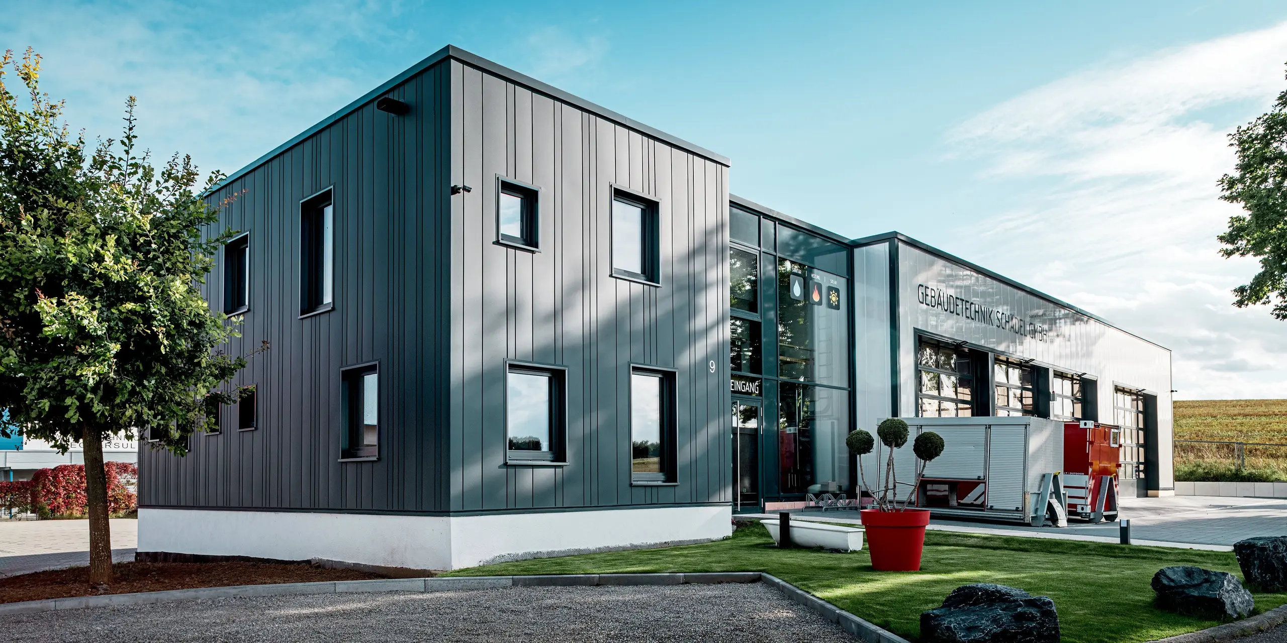 Die Szene zeigt den klar geformten Baukörper der Gebäudetechnik Schädel GmbH mit seiner prägnanten PREFA Blechfassade, deren vertikale Linien in Anthrazit sofort ins Auge fallen. Die Mischung aus verschiedenen Siding-Breiten verleiht dem Gebäude einen rhythmischen Charakter. Das Zusammenspiel aus Aluminium, Glasfront und offenem Vorplatz schafft eine moderne, kraftvolle Wirkung. Die langlebige PREFA Oberfläche bewährt sich gegen Wetter und Abnutzung.