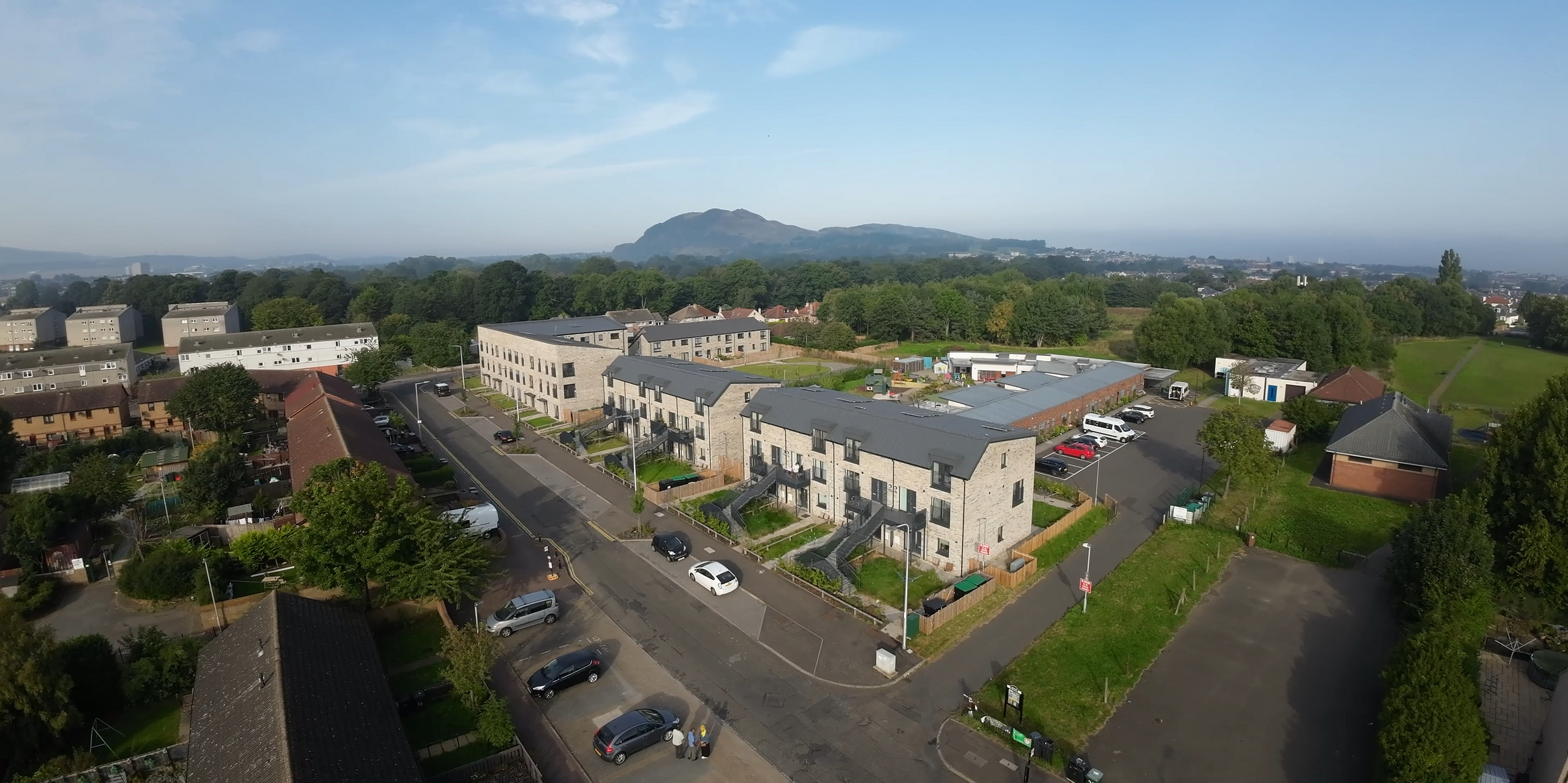 Aerial view of a residential complex in Bingham Avenue, Edinburgh. The residential complex is characterised by modern, two-storey buildings with PREFALZ roofs in P.10 dark grey. The roofs contrast with the light-coloured façades and blend harmoniously into the green, hilly landscape in the background. In the foreground you can see manicured green spaces and residential streets, while in the distance the striking Arthur's Seat, a well-known hill in Edinburgh, dominates the cityscape.