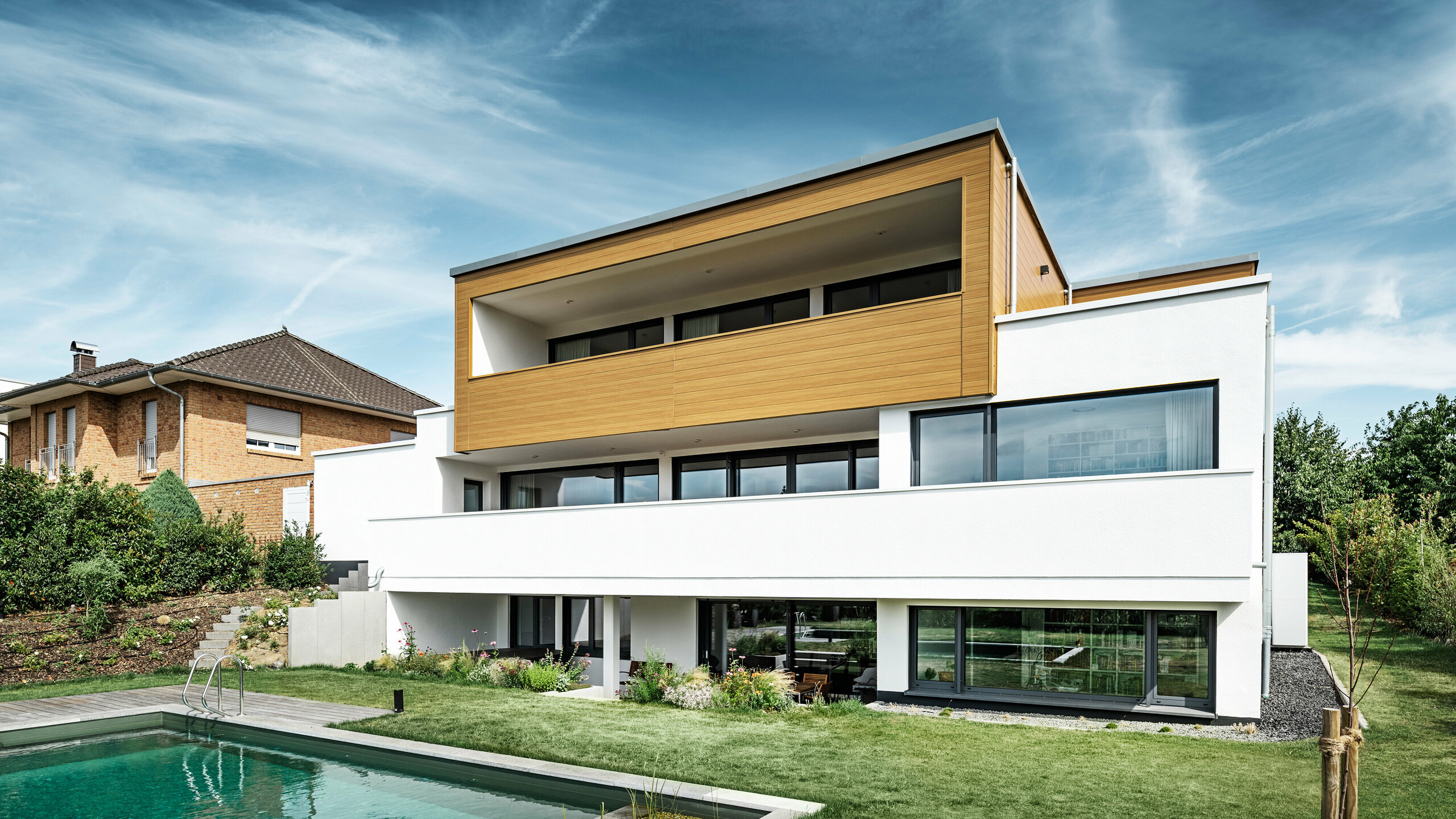 The rear of a modern detached house in Usingen, Germany. The green lawn, a pool and a young garden tree can be seen at the bottom of the picture. The eye-catching house has a white building base made of a rendered façade, an aluminium façade on the upper floor made of PREFA sidings in natural oak and large window areas with an anthracite-coloured frame. From a distance, the neighbouring house with a brick façade can be seen on the left edge of the picture.