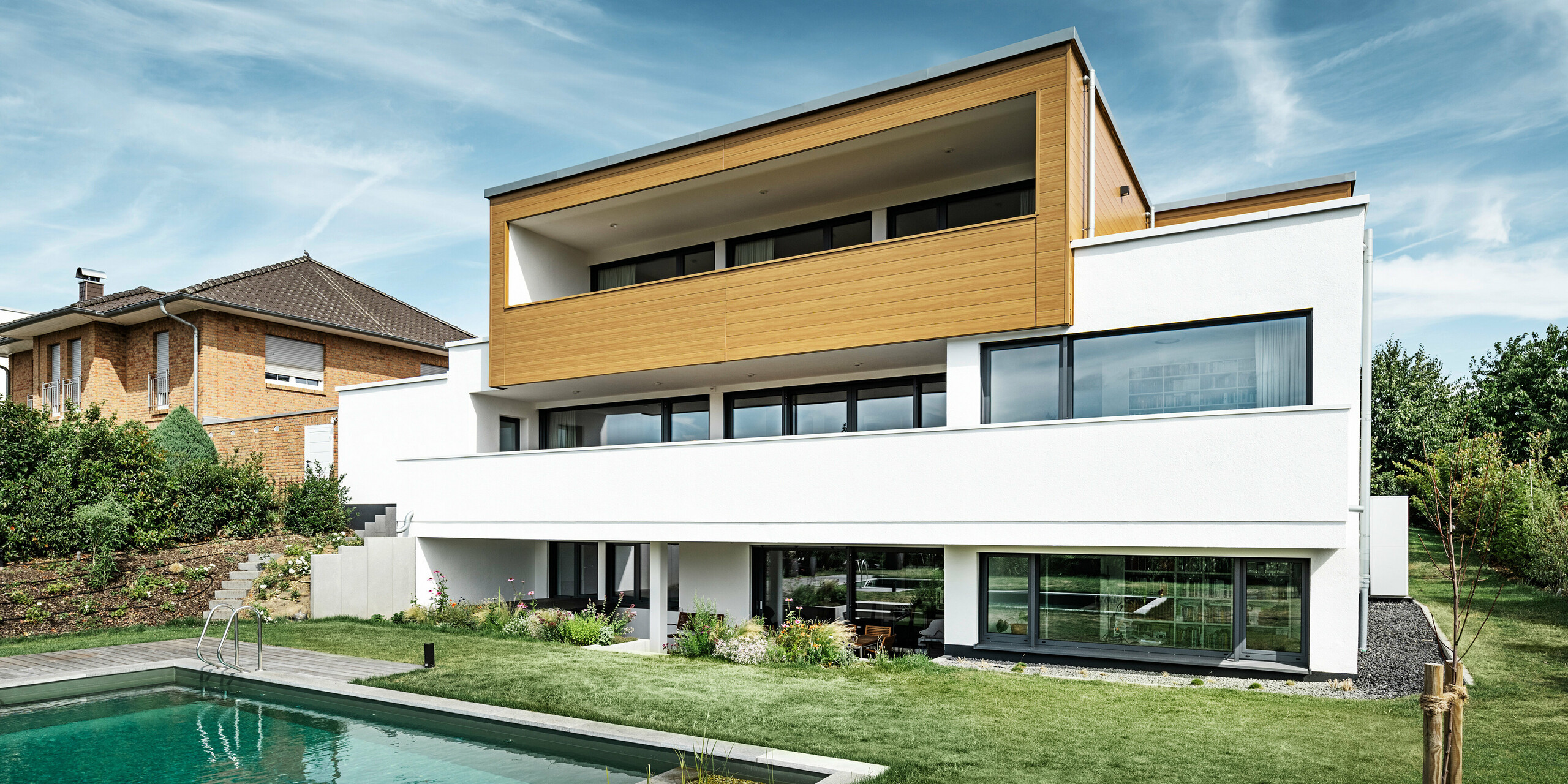 The rear of a modern detached house in Usingen, Germany. The green lawn, a pool and a young garden tree can be seen at the bottom of the picture. The eye-catching house has a white building base made of a rendered façade, an aluminium façade on the upper floor made of PREFA sidings in natural oak and large window areas with an anthracite-coloured frame. From a distance, the neighbouring house with a brick façade can be seen on the left edge of the picture.