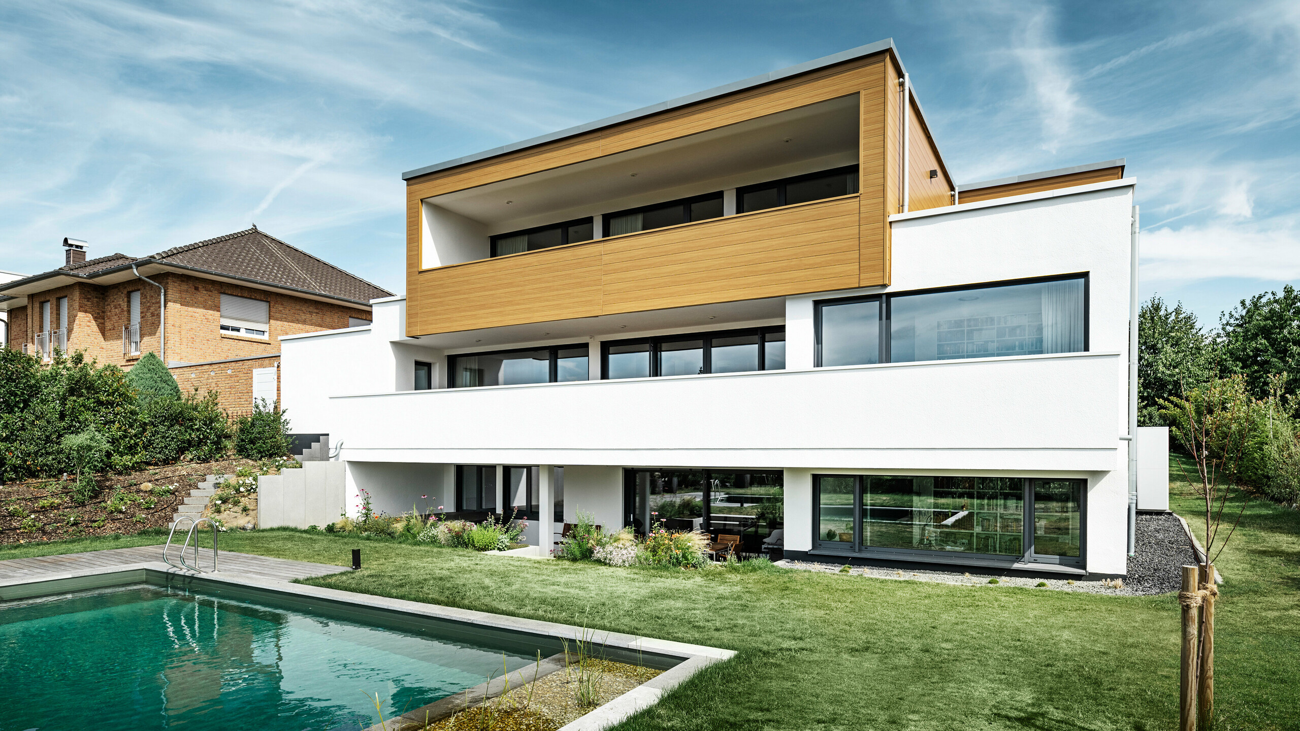 The rear of a modern detached house in Usingen, Germany. The green lawn, a pool and a young garden tree can be seen at the bottom of the picture. The eye-catching house has a white building base made of a rendered façade, an aluminium façade on the upper floor made of PREFA sidings in natural oak and large window areas with an anthracite-coloured frame. From a distance, the neighbouring house with a brick façade can be seen on the left edge of the picture.