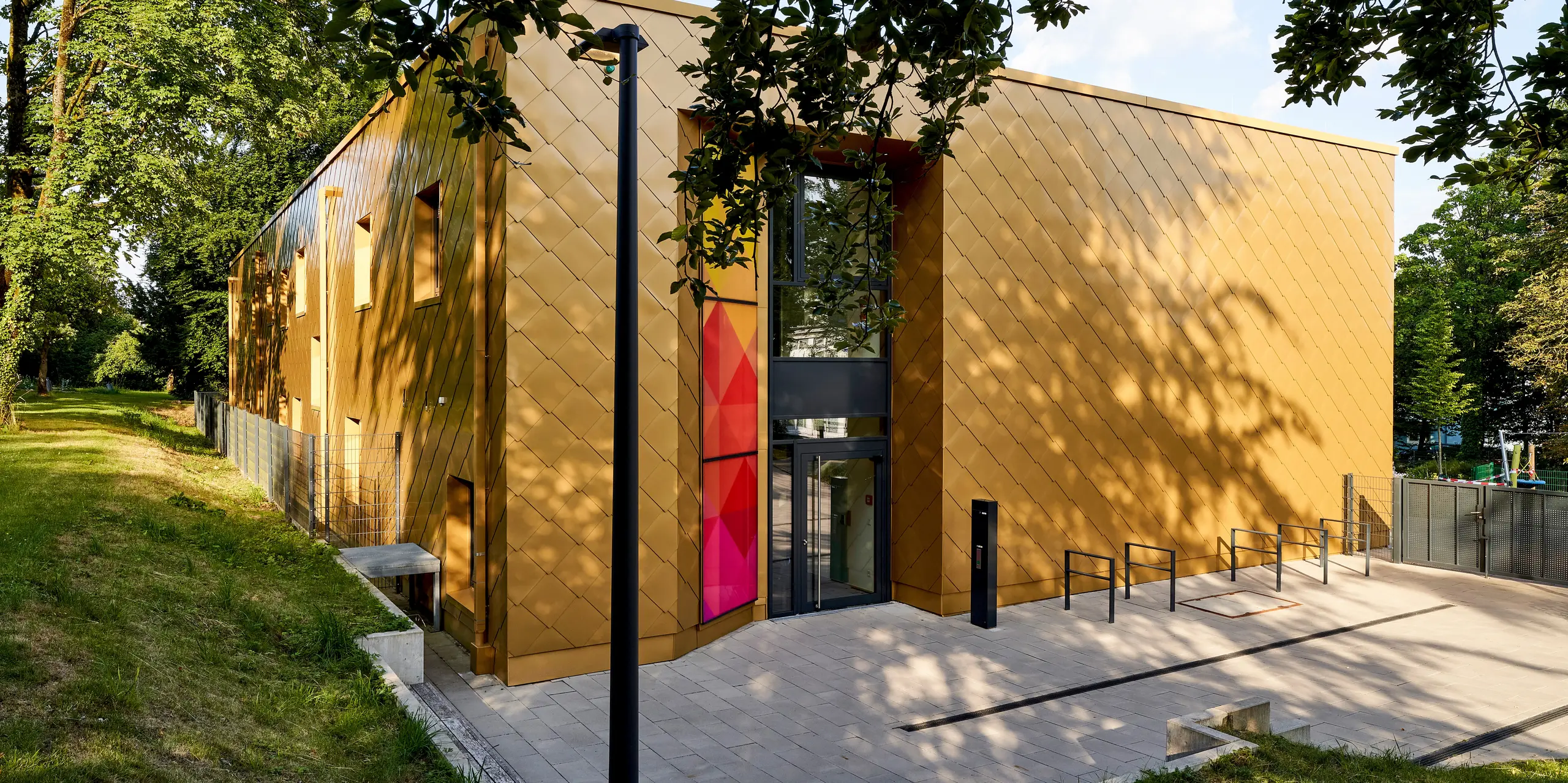 Außergewöhnliche Architektur mit goldfarbener PREFA Aluminiumfassade in präziser Wandrautenstruktur prägt diese eindrucksvolle Szene. Unter dem Blätterdach rahmen lebendige Licht- und Schattenspiele die markante Gebäudeform, während ein farbintensiver Eingangsbereich als echter Eyecatcher hervorsticht. Die diagonal verlegten Aluminiumrauten verleihen der Fassade Tiefe und Dynamik. PREFA Aluminiumprodukte überzeugen hier durch ihre Langlebigkeit, Witterungsbeständigkeit und brillante Farbqualität – eine nachhaltige, wartungsfreie Lösung für moderne Architektur im Einklang mit der Natur.