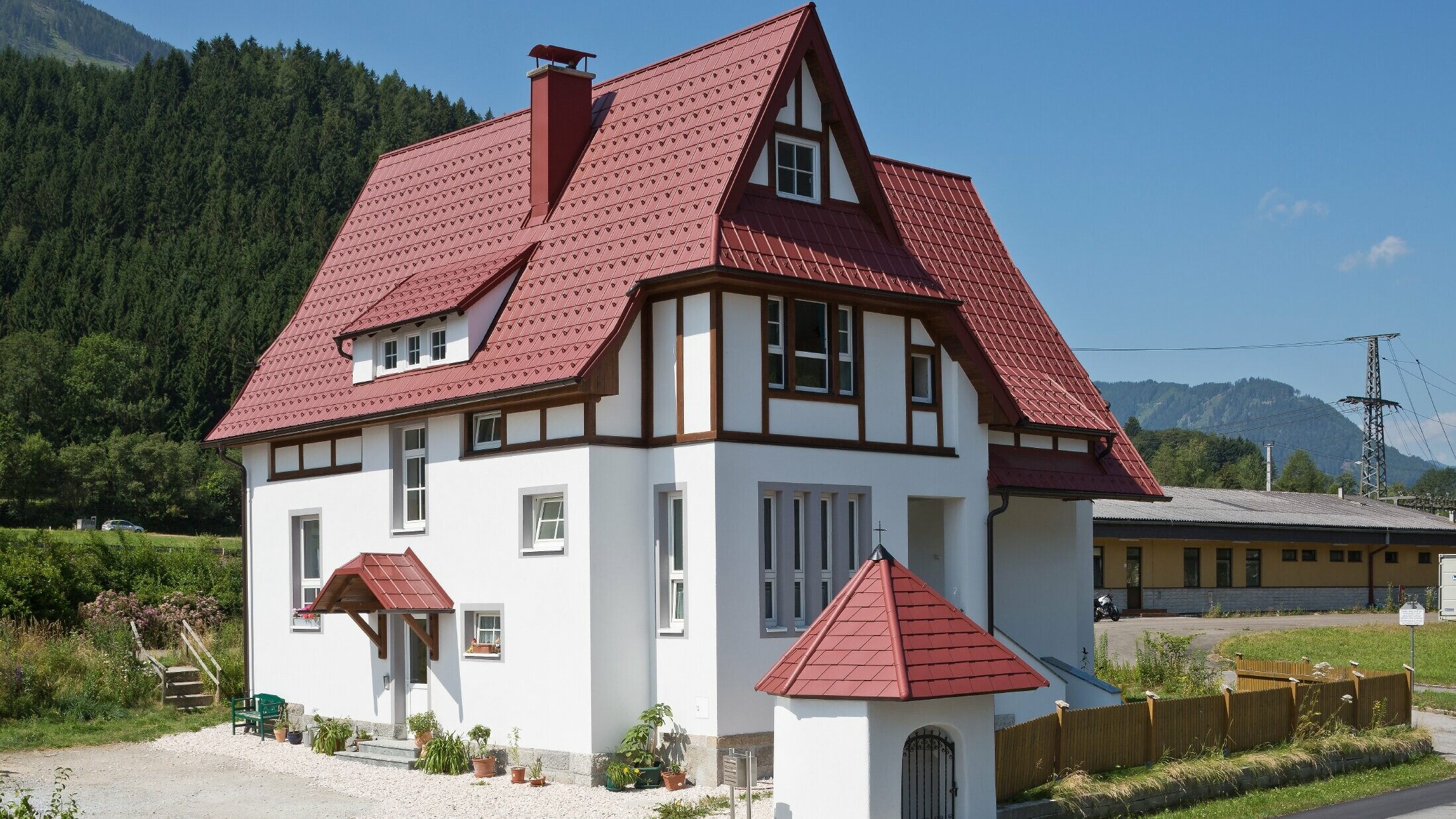 Steep, sloping roof with PREFA roof tiles in P.10 oxide red with many details such as dormers, turrets and chimney surrounds. The canopy and the wayside shrine in front of the house are also covered with PREFA sheet metal roofing.