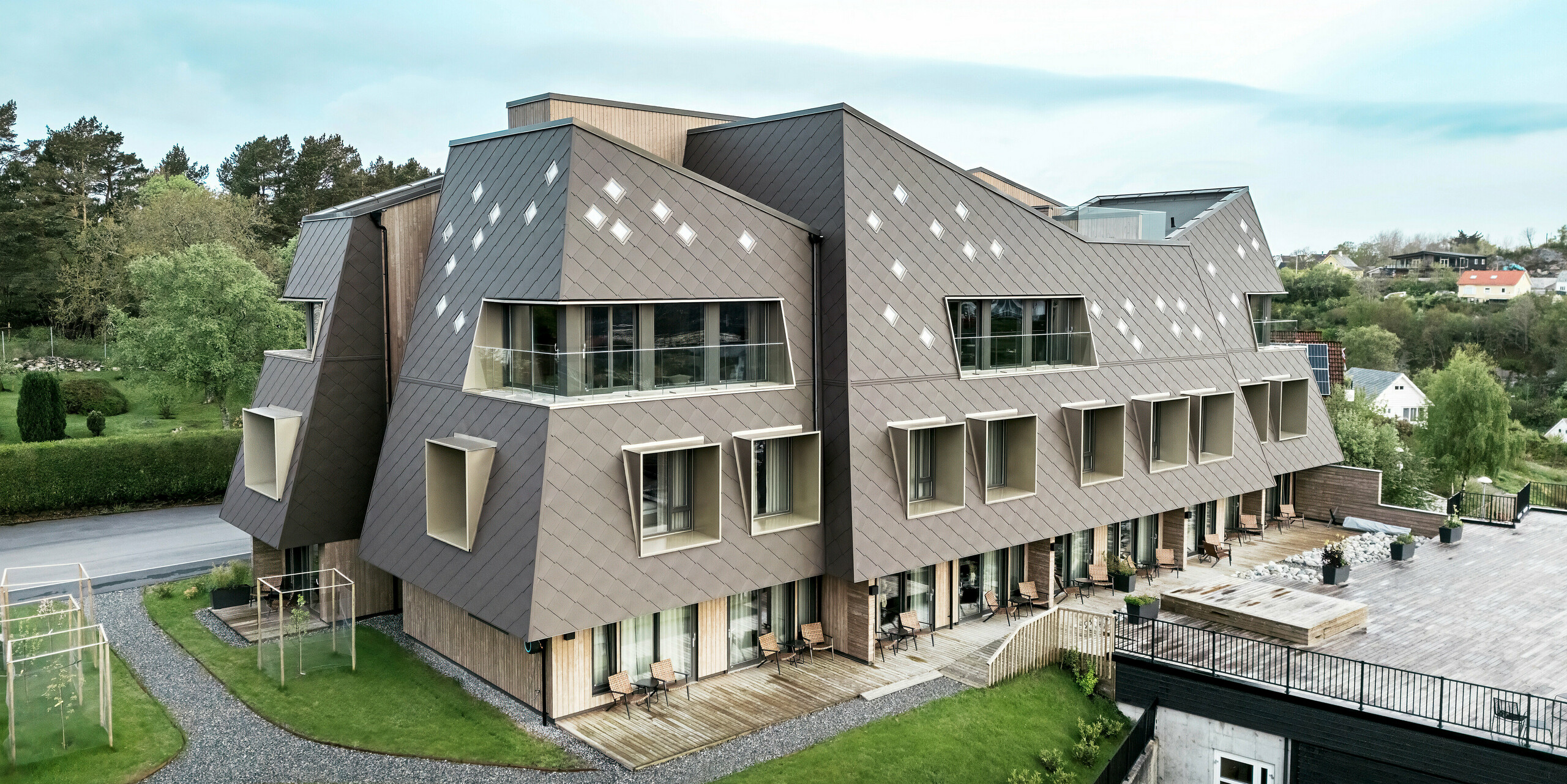 Außenansicht des Beckerwyc House in Bekkjarvik, Norwegen, mit Fokus auf die gepflegten Outdoor-Bereiche und die markante Fassade aus PREFA Wandrauten 44 × 44 in Nussbraun. Die skulpturale Architektur des Gebäudes, kombiniert mit einer großzügigen Terrasse und Balkonen, schafft eine harmonische Verbindung zwischen Innen- und Außenbereichen. Die futuristische Gestaltung und die natürliche Farbgebung passen sich gut in die umgebende Landschaft ein.