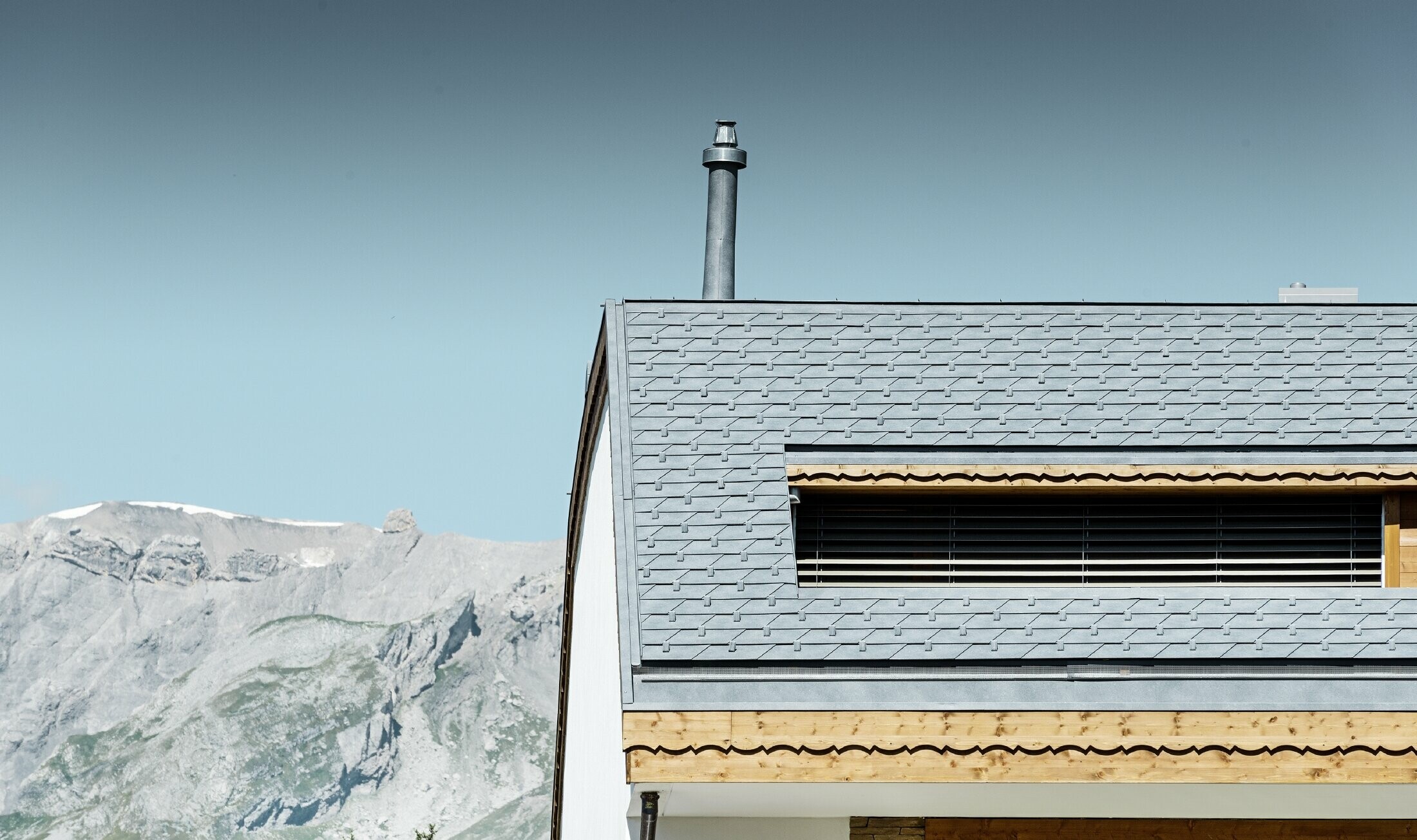 Apartment building in Crans-Montana (Switzerland) with mountains in the background, a façade with lovely wooden elements and a PREFA aluminium shingle roof in stone grey