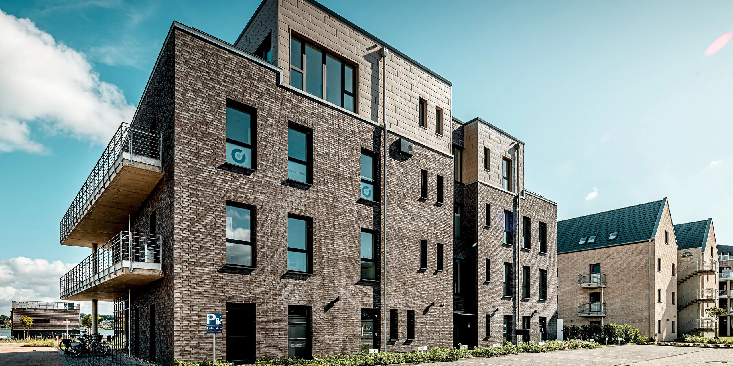 Das Bild zeigt ein Gebäude der Wohnanlage „Hafen zur Freiheit“, das durch die Kombination aus dunkler Klinkerfassade und den markanten PREFA Siding.X Paneelen in Bronze im Staffelgeschoss besonders modern wirkt. Die klar strukturierten Aluminiumflächen setzen einen eleganten Kontrast zur traditionellen Ziegeloptik und verleihen dem Baukörper eine dynamische, hochwertige Ausstrahlung. Großzügige Fenster und seitliche Balkonflächen betonen die offene Architektur. Dank der langlebigen, witterungsfesten PREFA Elemente bleibt die Fassade auch in der maritimen Umgebung dauerhaft geschützt und optisch beständig.