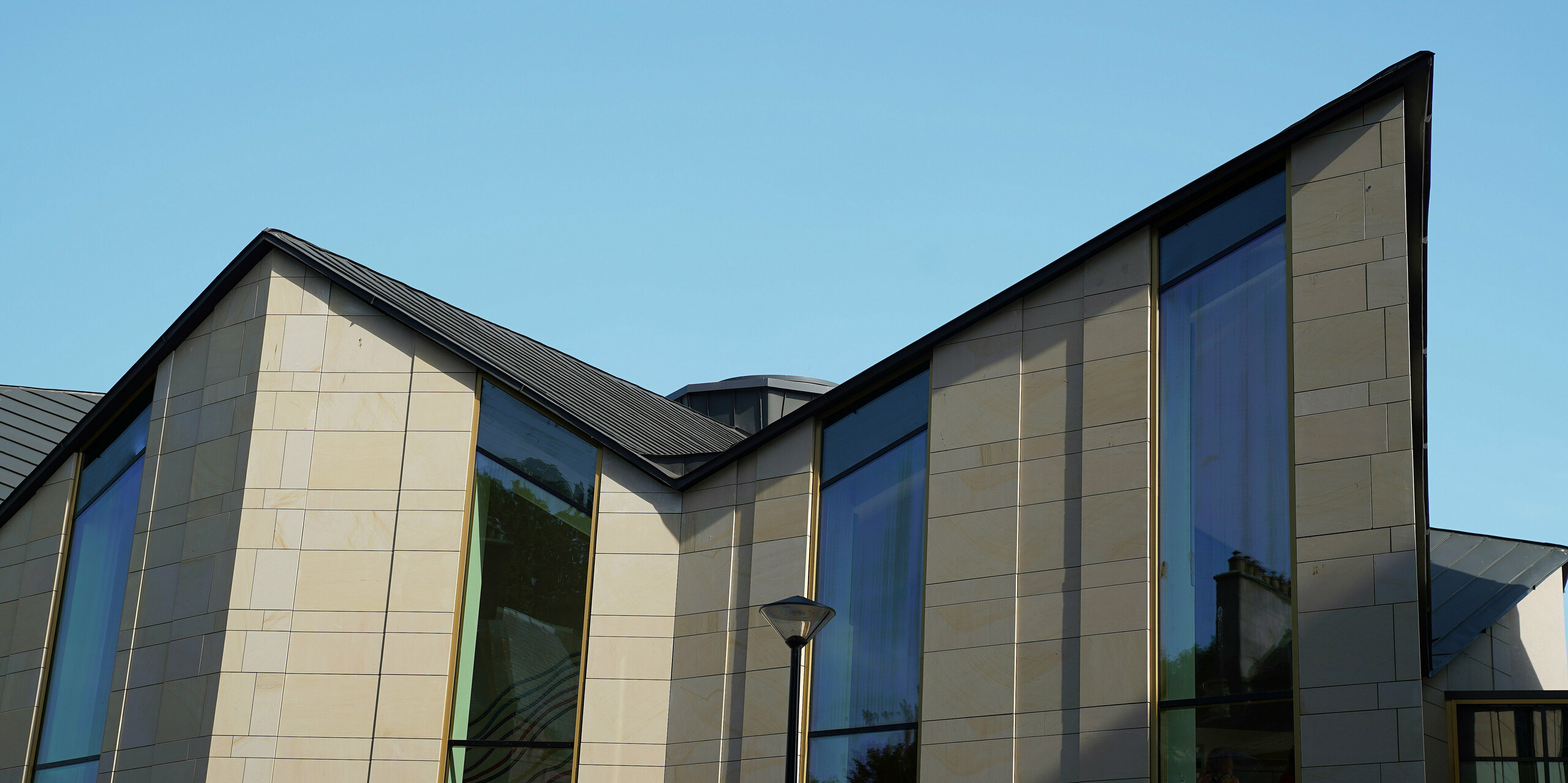 Architectural detail shot of the roof of 'The Great Tapestry of Scotland' visitor centre in Galashiels. The clean lines of the PREFALZ roof in P.10 zinc grey form an impressive contrast to the large, light-coloured stone façade and the generous glass fronts. The roof construction demonstrates the flexibility and aesthetics of the PREFALZ roof system, which combines modern design with functional excellence.