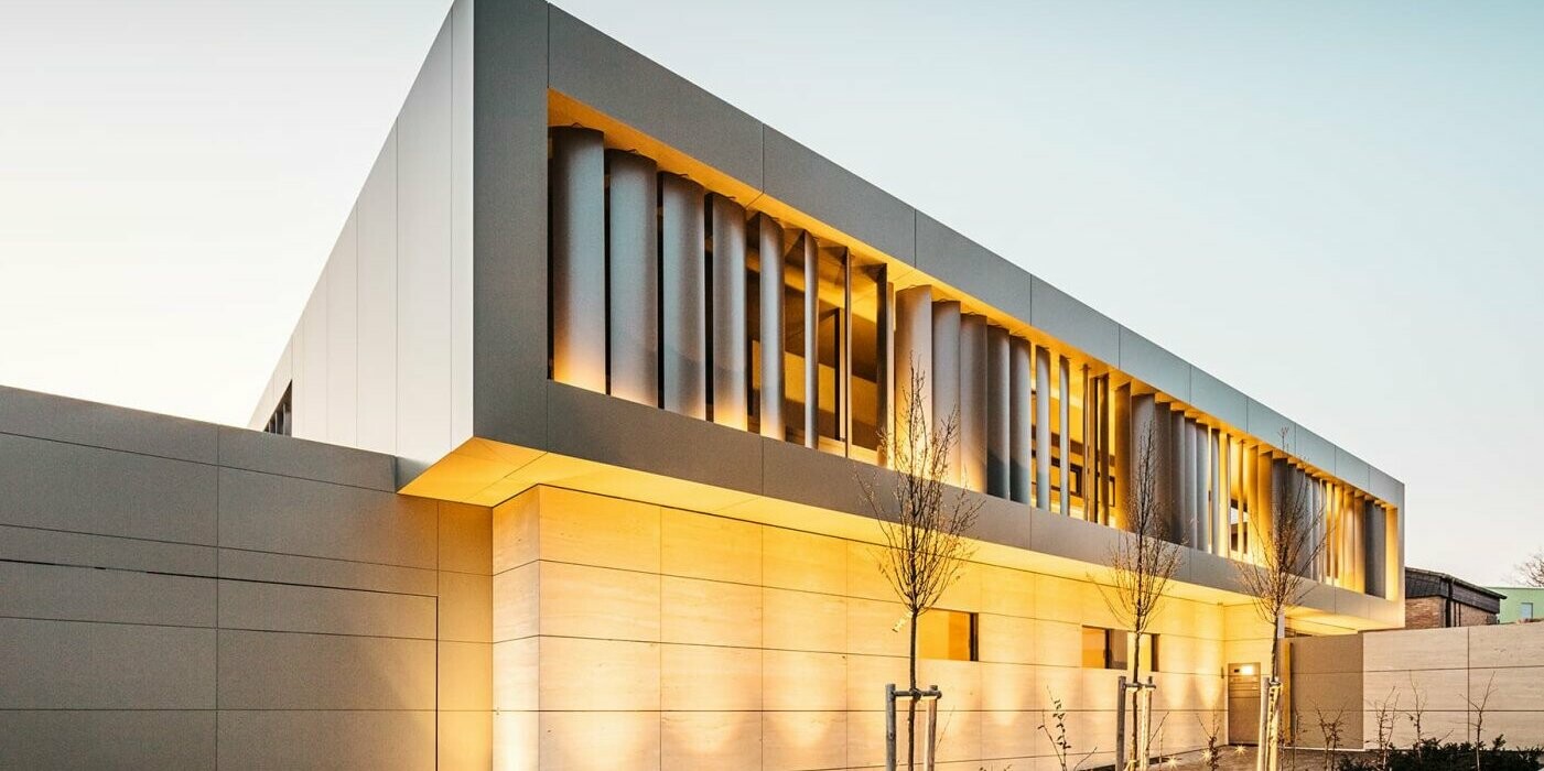 Street view at night, garage with bronze-coloured façade on the left, stone façade below, bronze-coloured aluminium façade with slats inside above