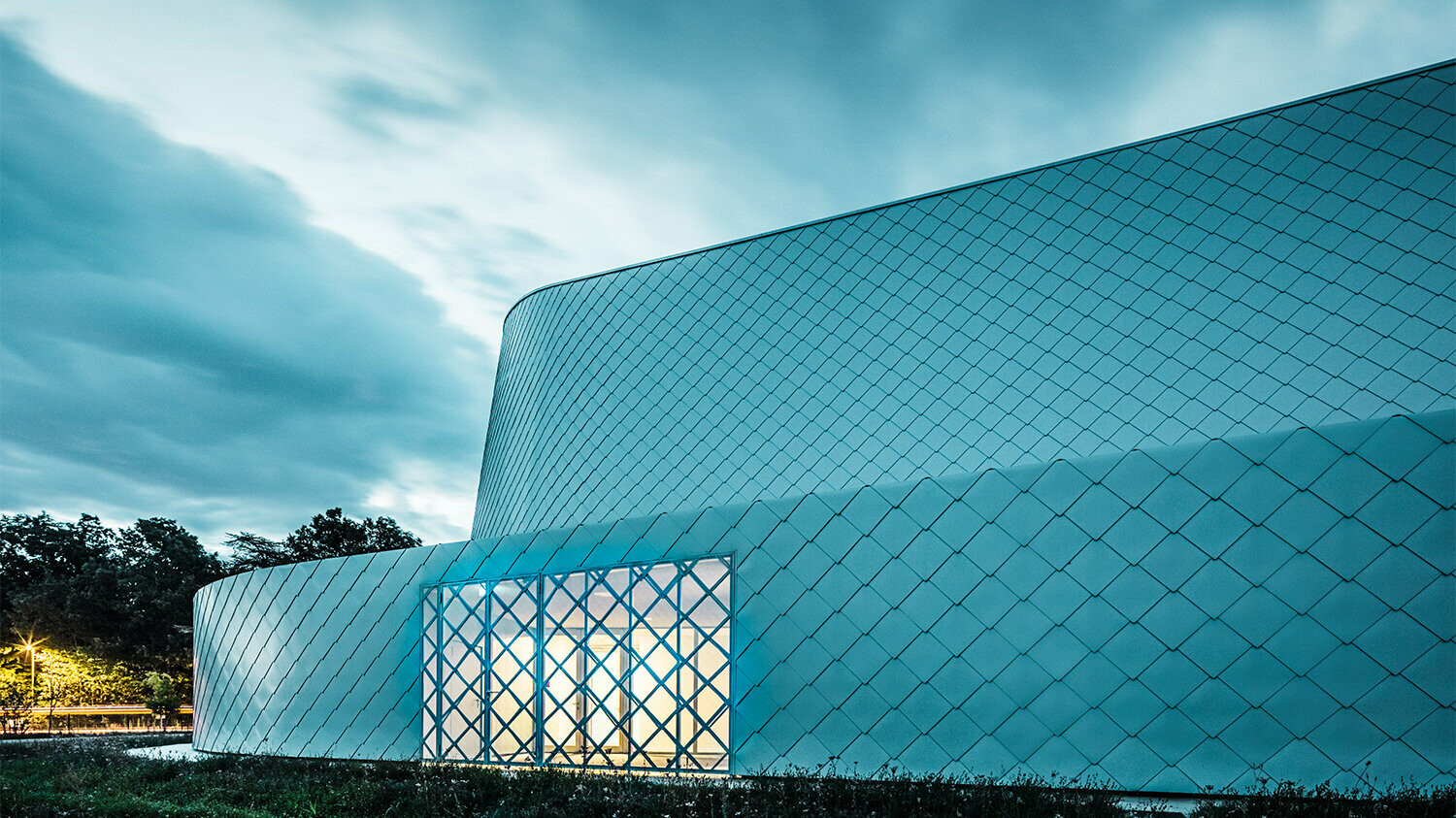 Lateral evening evening shot of the illuminated building, the window is barred with the same patterns as that of the rhomboid tile façade.
