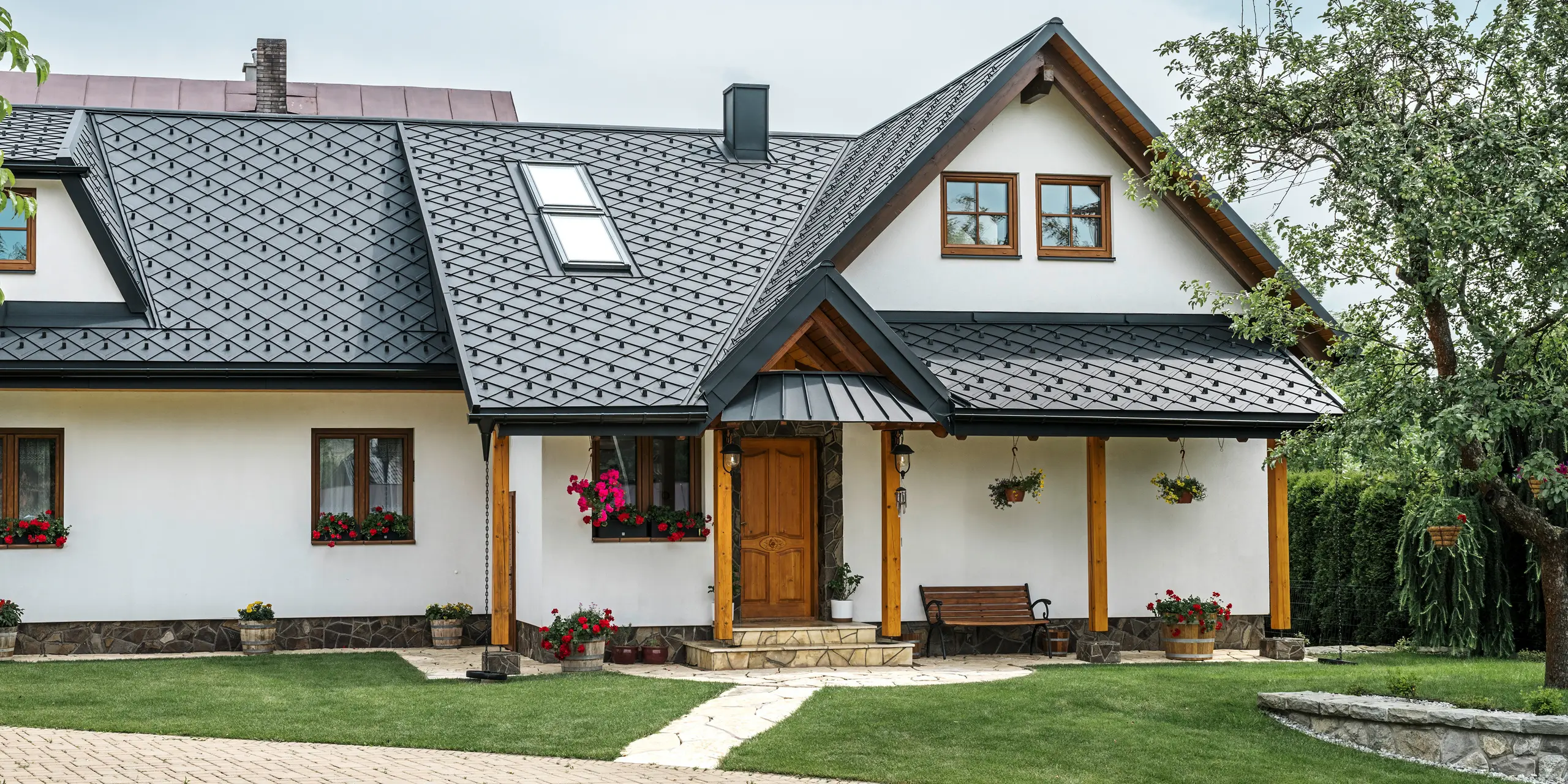 Eingangsbereich eines traditionellen Einfamilienhauses in Stara Bystrica, Slowakei, mit einem Aluminiumdach aus PREFA Dachrauten 29 × 29 in P.10 Schwarz. Das hochwertige Blechdach in eleganter Schuppenoptik umfasst Dachschrägenfenster und eine Veranda mit Vordach, das mit PREFALZ verkleidet ist. Die eleganten und zugleich robusten Aluminiumprodukte verleihen dem Haus ein zeitgemäßes und einladendes Erscheinungsbild. Der gepflegte Garten und die Blumenarrangements unterstreichen die gute Pflege des Gebäudes.