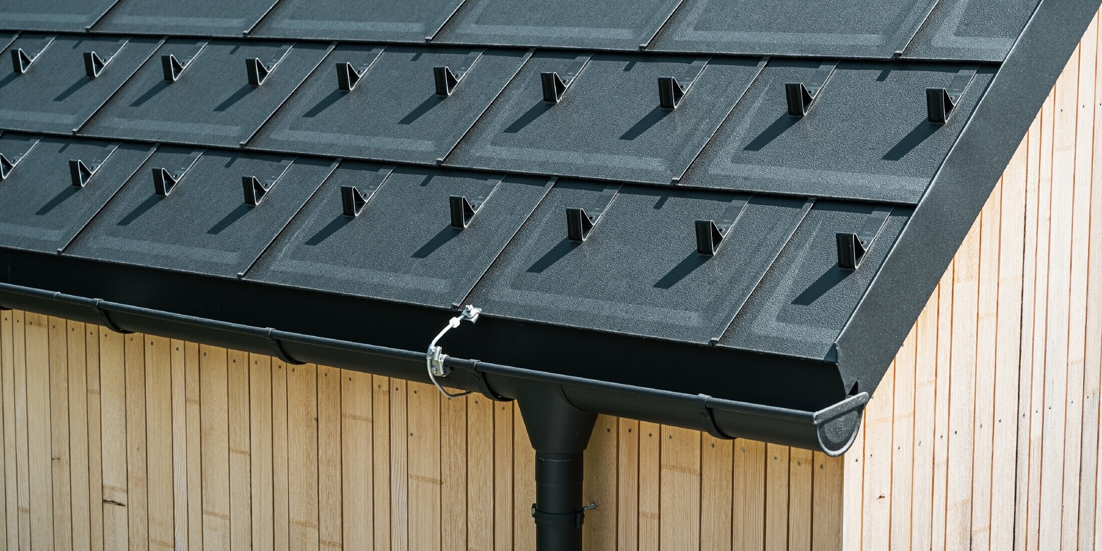 Close-up of PREFA roof tile R.16 with snow guards, gutter and downpipe in black P.10 incl. lightning protection and a vertical wooden façade