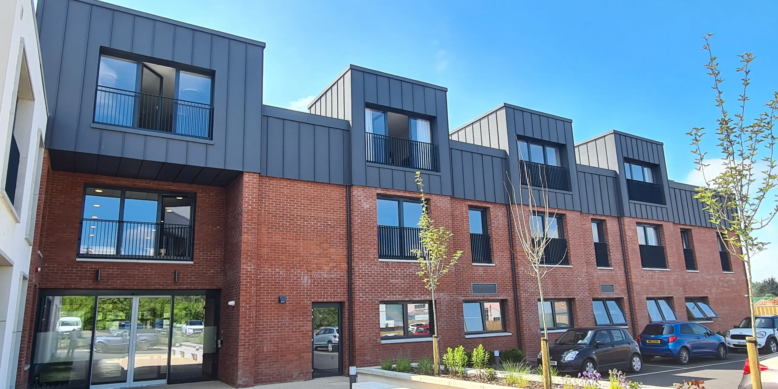 Front view of Harberton Hall in Belfast, Northern Ireland. The standing seam roof made of PREFALZ P.10 dark grey perfectly complements the brick façade. Large windows and flat roof dormers emphasise the innovative design of the building for assisted living and create a welcoming atmosphere. The glass entrance door provides insights into the internal facilities.