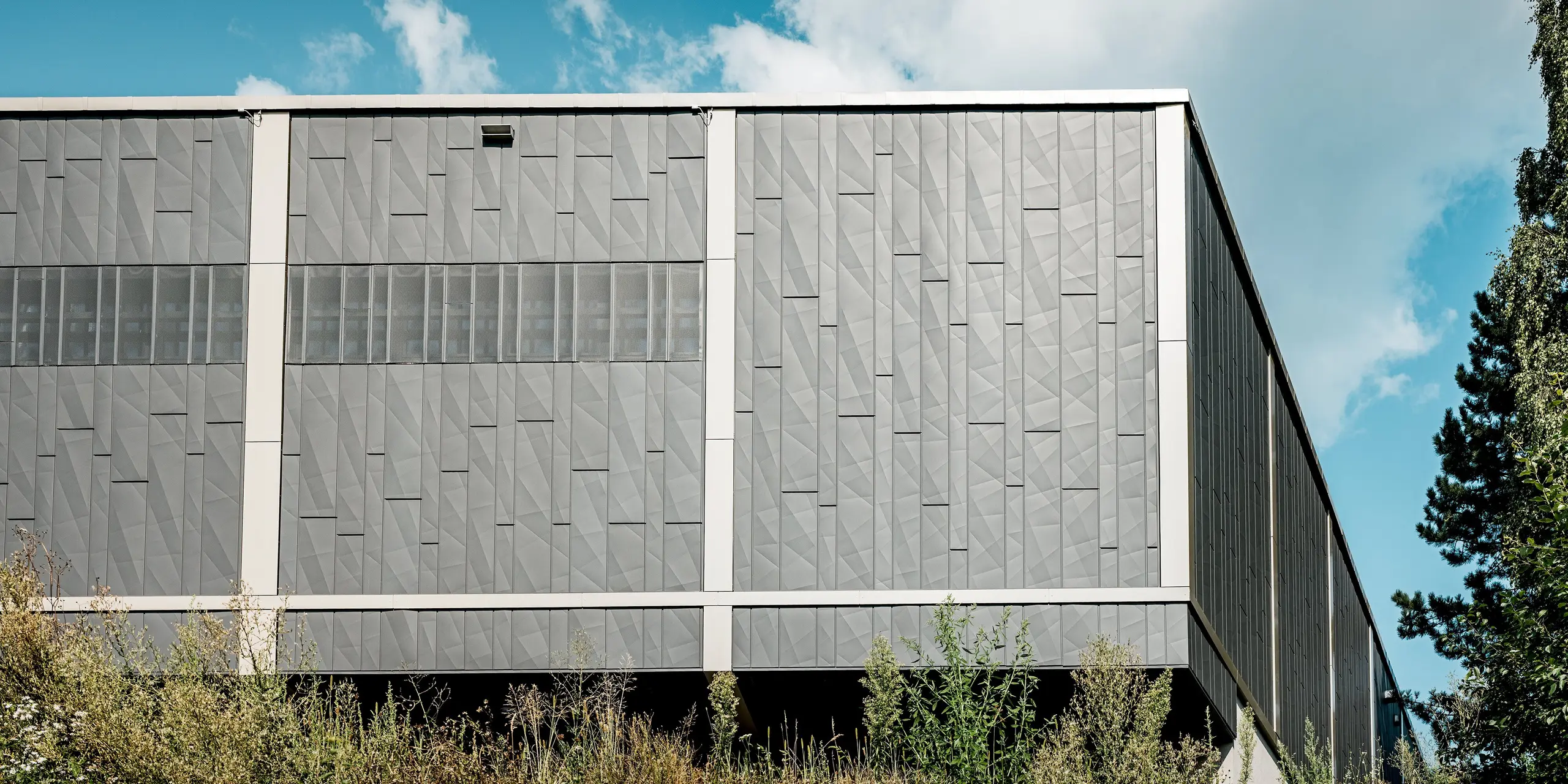 Das Bild zeigt die Rückseite des Einkaufszentrums in Rohrbach mit einer modernen Blechfassade aus PREFA Siding.X-Paneelen in Anthrazit. Die geometrischen Strukturen der Paneele schaffen eine dynamische Textur, die dem Gebäude eine markante und zeitgenössische Ästhetik verleiht. Besonders auffällig sind die horizontalen Streifen aus perforierten Elementen, die nicht nur eine optische Vielfalt bieten, sondern auch für eine gezielte Belüftung und Lichtdurchlässigkeit sorgen. Die Fassade fügt sich harmonisch in die umgebende Natur ein, wobei das Spiel zwischen den satten grauen Tönen und dem grünen Bewuchs eine reizvolle Verbindung zwischen Architektur und Landschaft bildet.