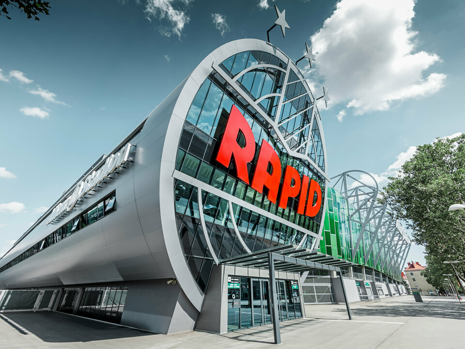 Ansicht des Allianz Stadions von SK Rapid Wien mit auffälliger PREFA Fassade in Form der Aluminium Verbundplatte in Silbermetallic. Die Gebäudehülle strahlt Dynamik und Moderne aus. Der Eingangsbereich, gekrönt mit dem leuchtend roten RAPID-Schriftzug, lädt ein in das Herz des Fußballs, eingefasst von einer futuristischen und zugleich eleganten Fassadenstruktur, die Sportarchitektur auf ein neues Niveau hebt.