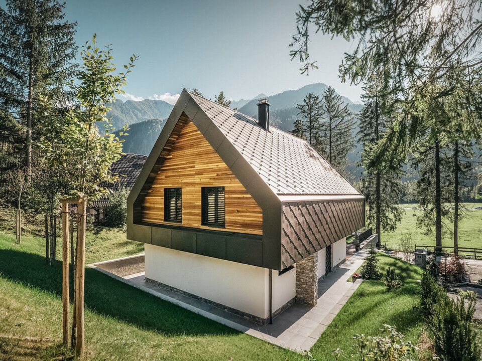 A modern chalet in Slovenia with a combination of traditional and contemporary architectural elements, embedded in an alpine landscape. The house features a wooden façade with closed shutters, a characteristic steep pitched roof with nut-brown roof and wall rhombuses and a chimney. The property is surrounded by abundant green spaces, trees and offers a clear view of the distant mountains and an open valley