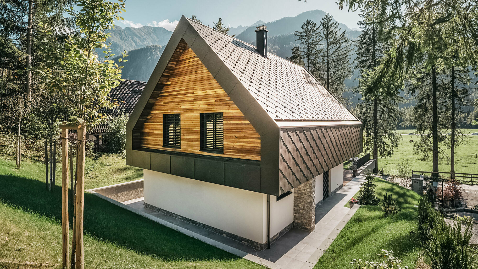 A modern chalet in Slovenia with a combination of traditional and contemporary architectural elements, embedded in an alpine landscape. The house features a wooden façade with closed shutters, a characteristic steep pitched roof with nut-brown roof and wall rhombuses and a chimney. The property is surrounded by abundant green spaces, trees and offers a clear view of the distant mountains and an open valley