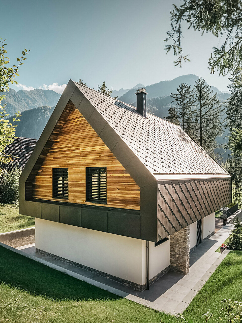 A modern chalet in Slovenia with a combination of traditional and contemporary architectural elements, embedded in an alpine landscape. The house features a wooden façade with closed shutters, a characteristic steep pitched roof with nut-brown roof and wall rhombuses and a chimney. The property is surrounded by abundant green spaces, trees and offers a clear view of the distant mountains and an open valley