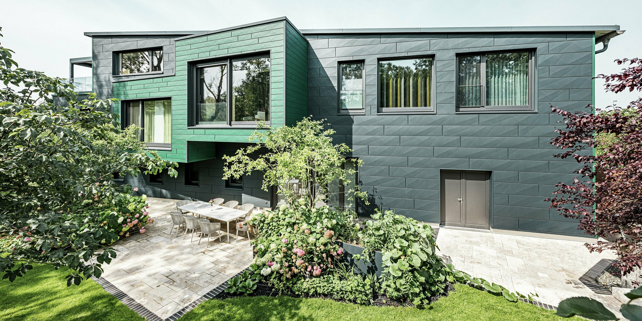 The detached house in Türkheim impresses with its unusual façade, which is clad with PREFA Siding X panels in anthracite and moss green. The clean lines and striking colour choice of the aluminium panels give the building a modern and elegant look. In the foreground is the spacious garden area with a stylish terrace and a dining table with modern chairs. Colourful flower beds and healthy plants provide a lively, green contrast to the robust façade. The durable and low-maintenance aluminium façade protects the building and ensures timeless beauty.